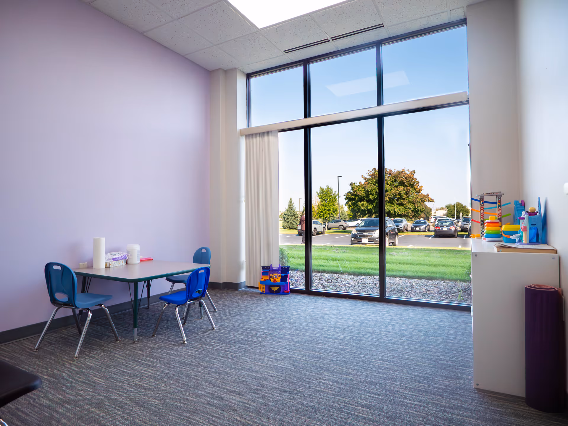 Small playroom with purple walls, large windows, a table and blue chairs, plus toys.