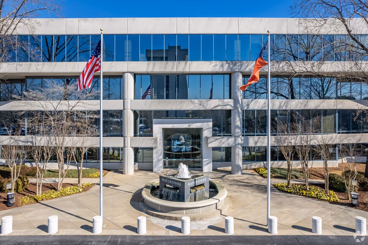 Exterior view of a modern facility entrance with glass facade, flags, and a circular fountain.
