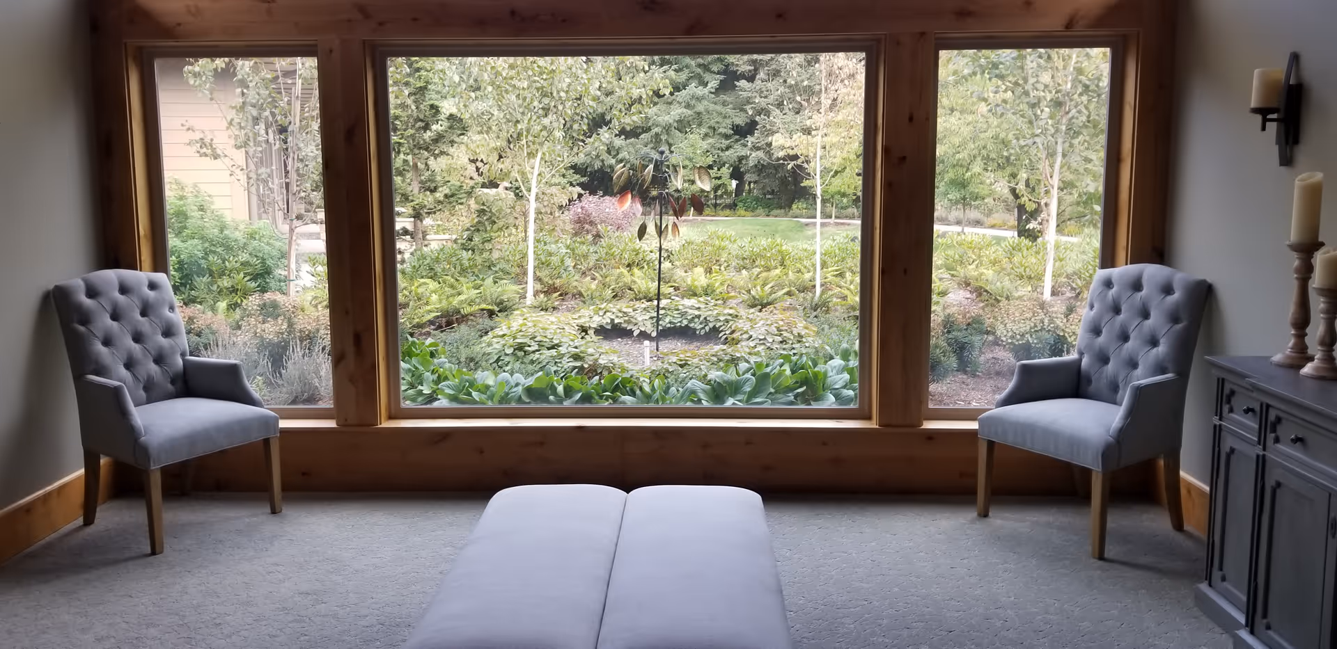 Cozy living room with two gray tufted chairs facing a large window overlooking a garden.