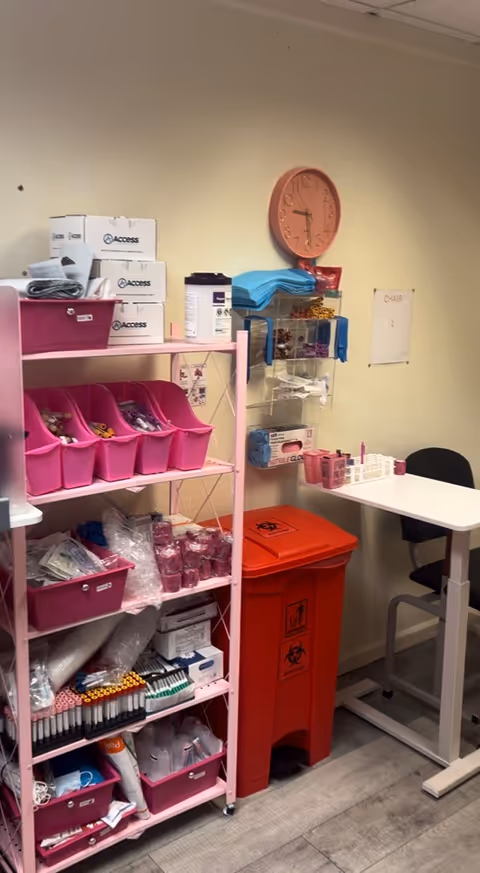 Medical supply closet with pink shelves, boxes, and orange biohazard bin beside a table.