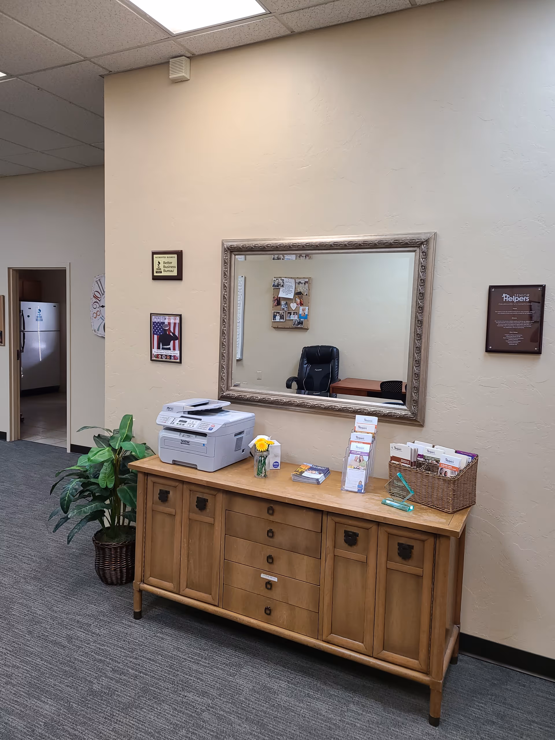 Reception area with wooden cabinet, printer, brochures, mirror, and plant in a neutral hallway.