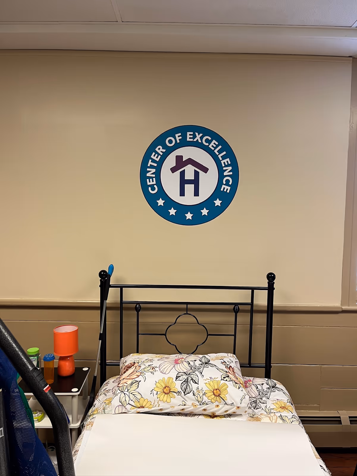 Hospital-style bedroom with metal headboard, floral bedding, and a Center of Excellence wall logo.