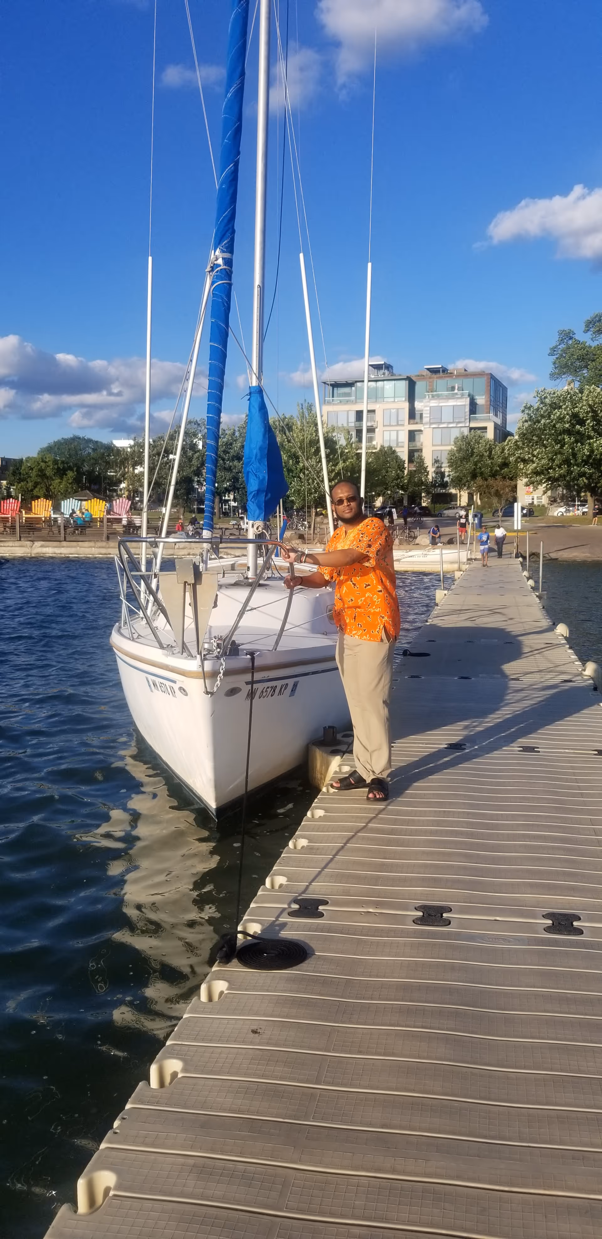 A man stands on a marina dock beside a sailboat under a blue sky.