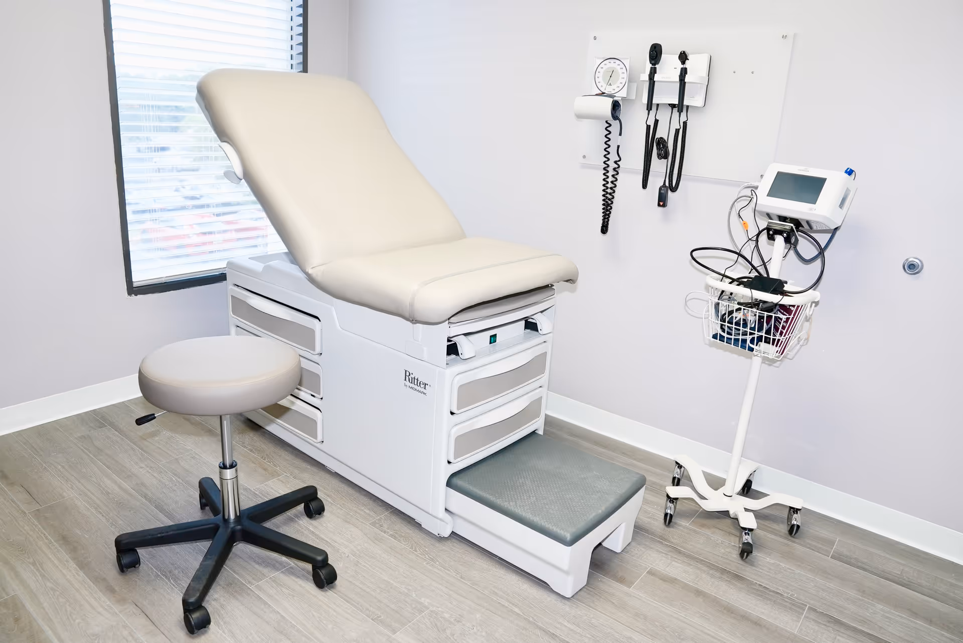 A clean medical examination room with a reclining chair, stool, and mobile monitoring cart.