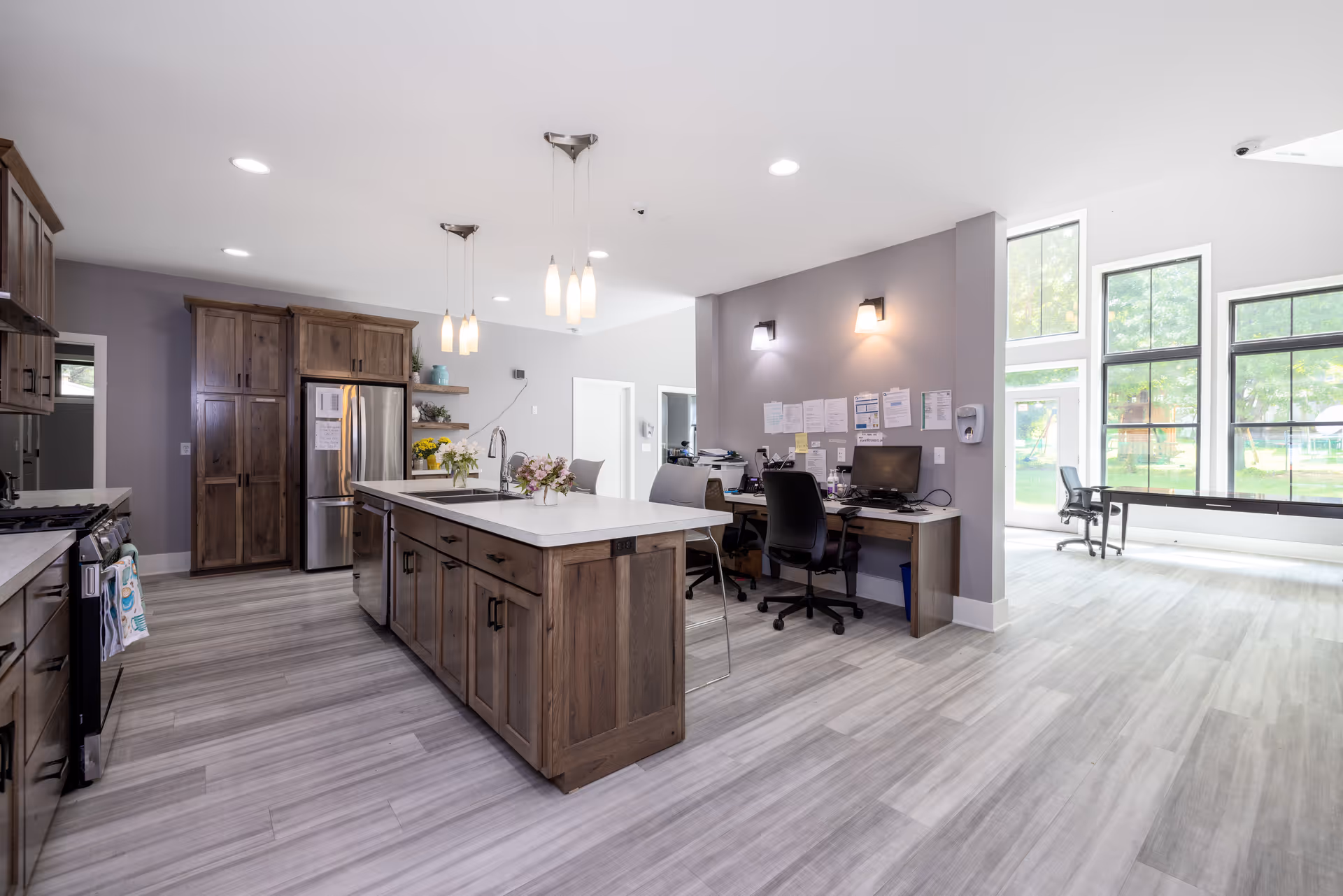 Bright communal kitchen with island, wooden cabinets, and a desk area by large windows.
