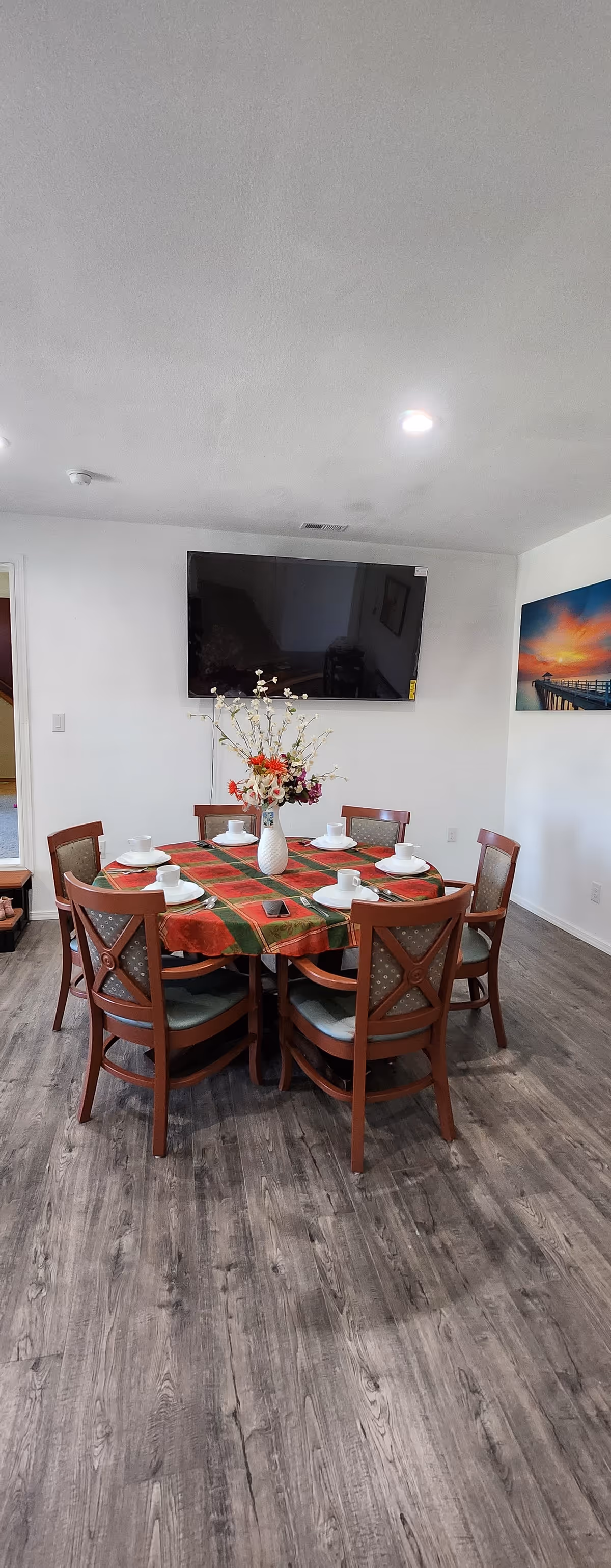 A round dining table with six chairs, plates, flowers, and a wall-mounted TV.