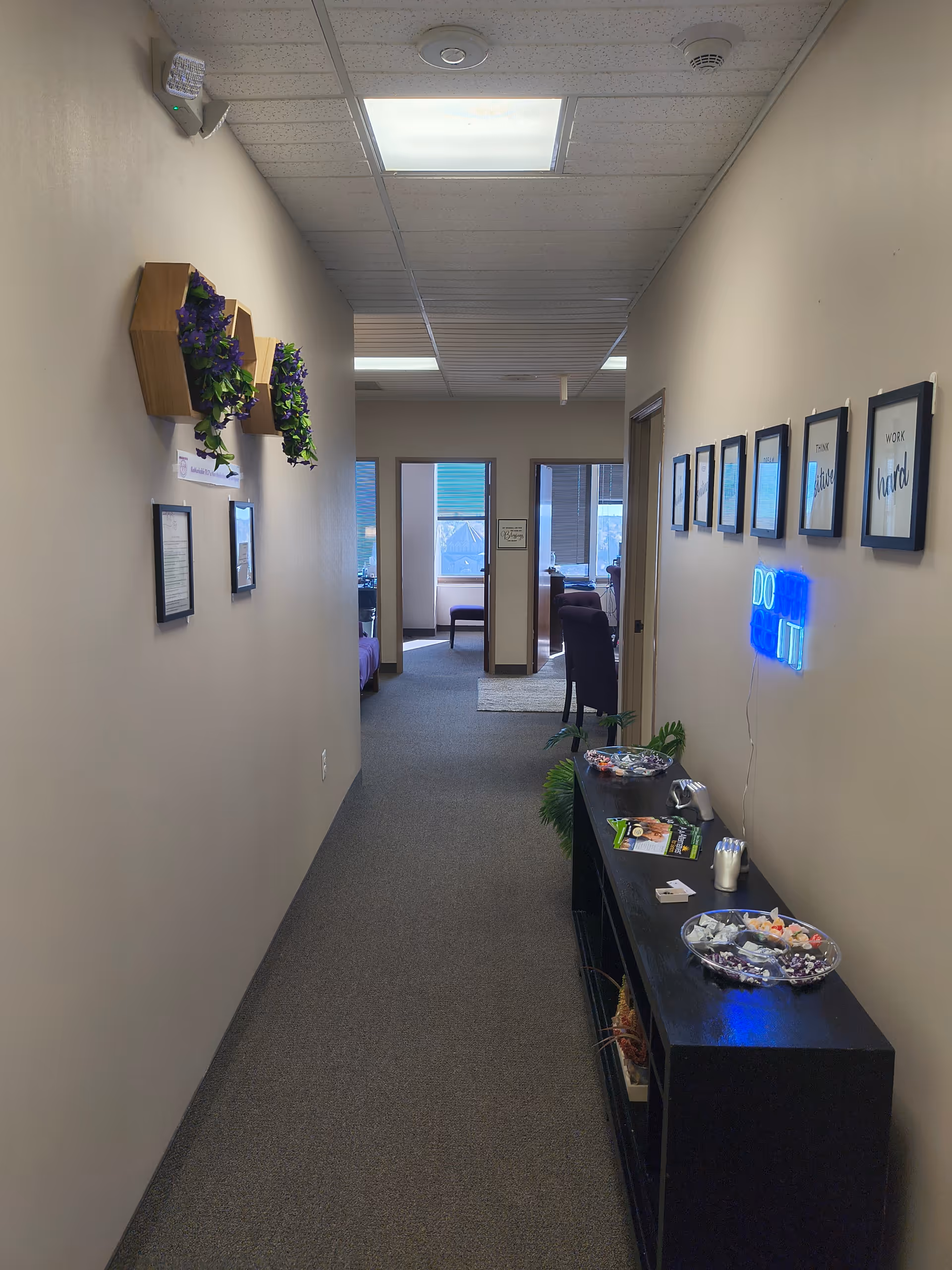 Narrow beige hallway in a senior facility with framed art, plants, and a black sideboard.