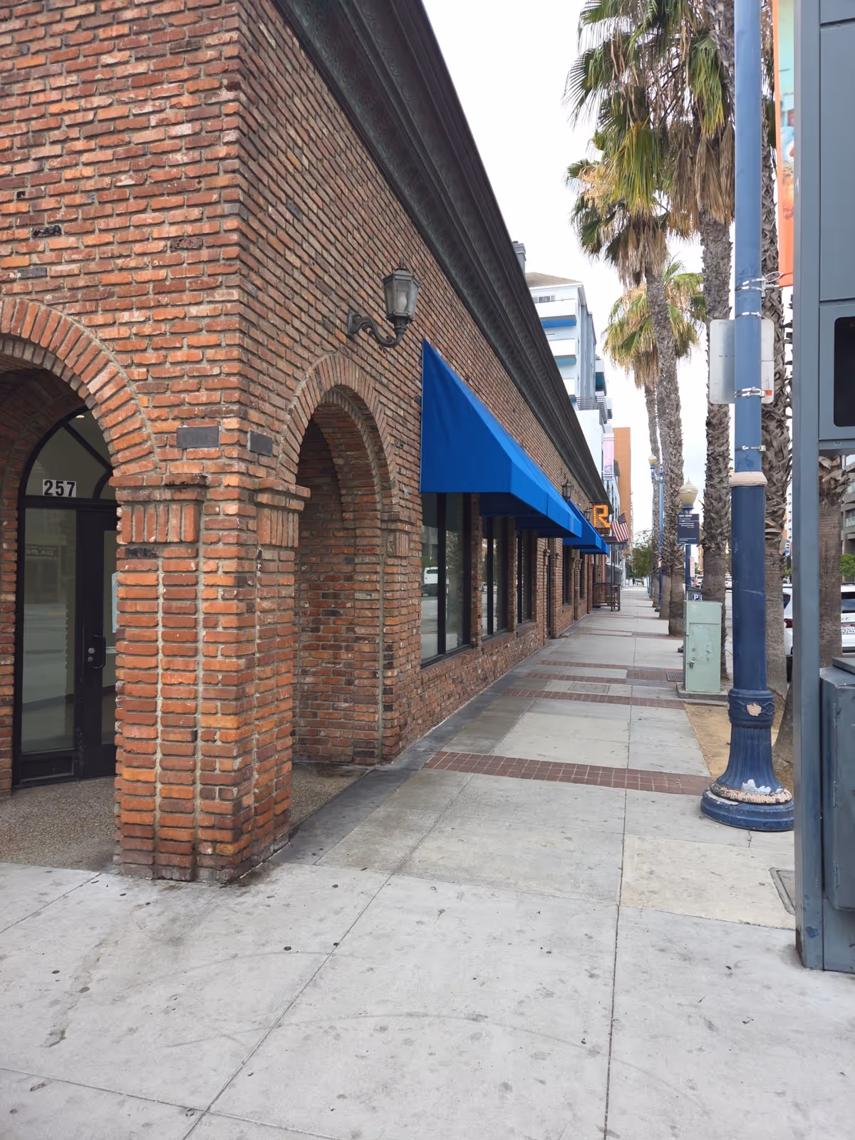 Brick storefronts line a sidewalk with arched entrances, blue awnings, and palm trees.