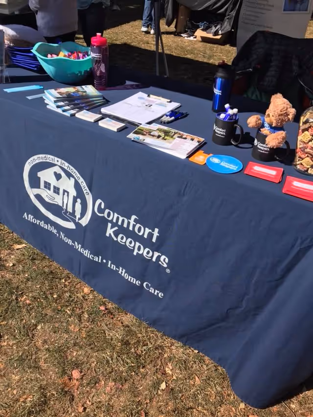 An outdoor booth displays a Comfort Keepers Home Care tablecloth, brochures, and promotional items.