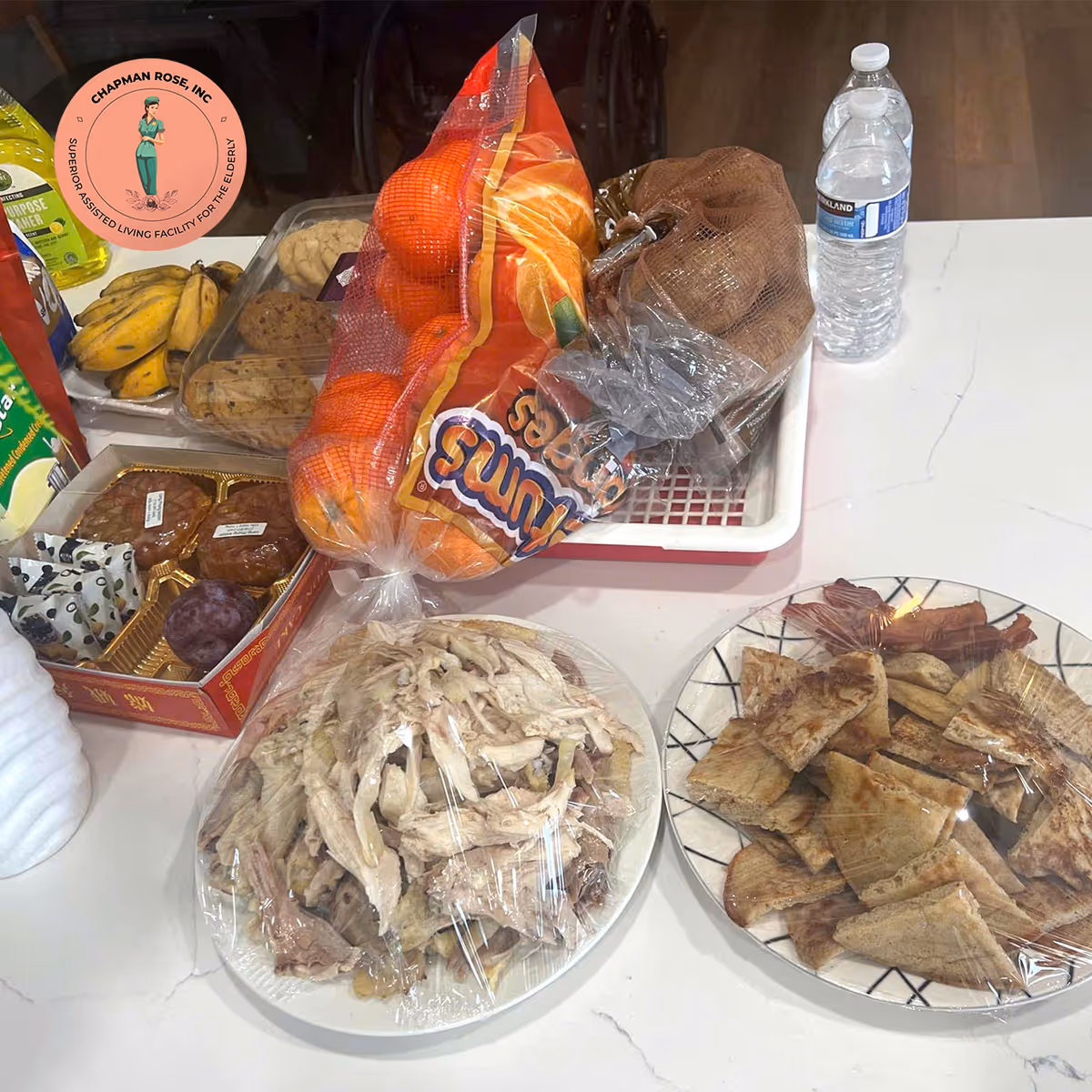 Assorted foods on a counter, including fruit, bread, chips, and sliced meat plates.