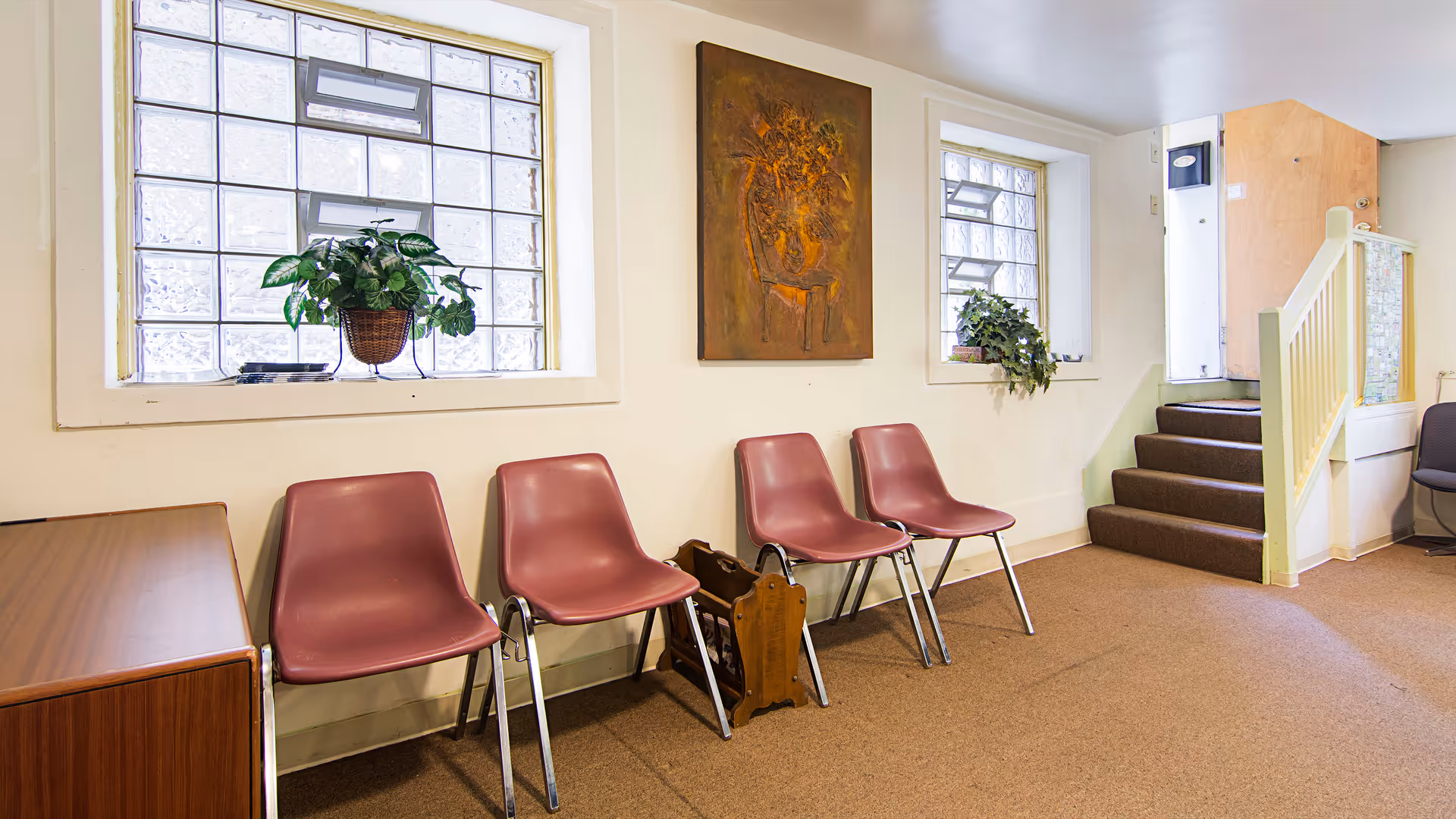 A hallway waiting area with pink chairs, windows, plants, artwork, and a staircase.