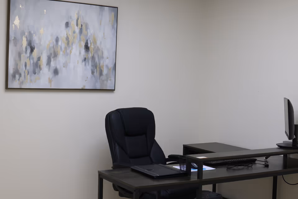 A dark desk with laptop, monitor, chair, and abstract painting on the wall.