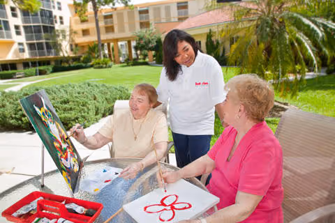Three people outdoors, two elderly residents painting while a caregiver assists beside them.