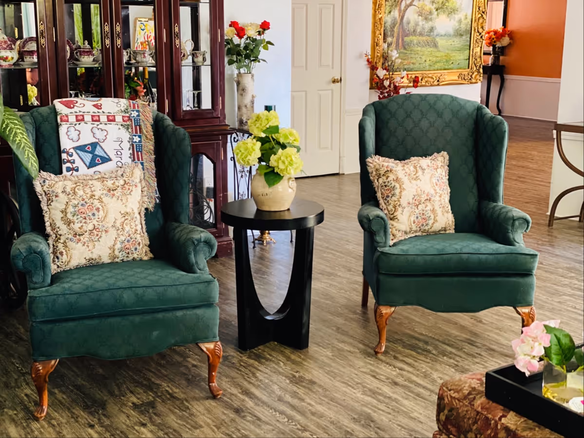 Two green upholstered armchairs with pillows sit in a living area beside a table and flowers.