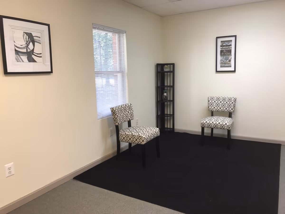 A small, empty waiting area with two patterned chairs, a tall shelving unit, and wall art.