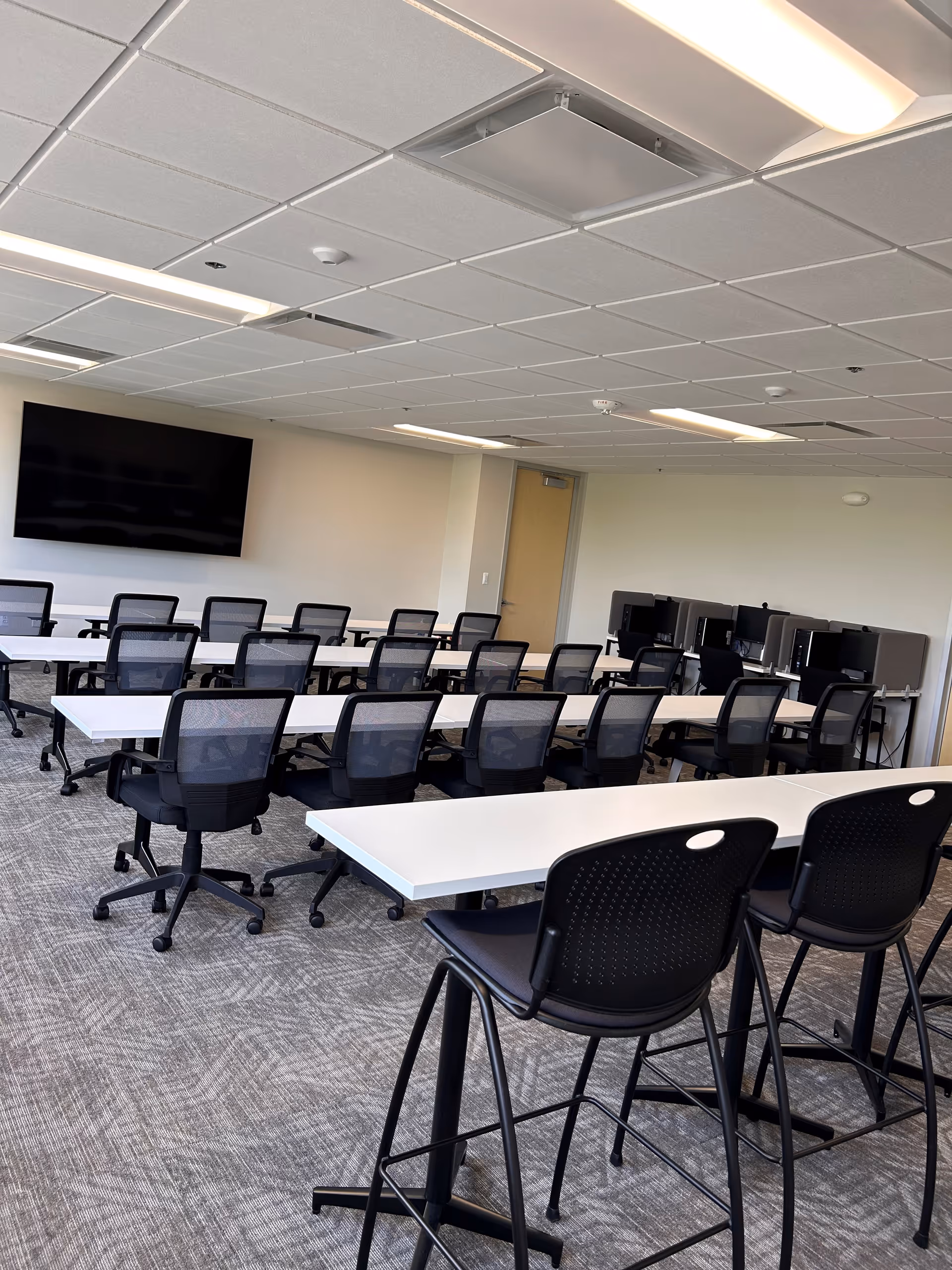 Modern computer lab or training room with desks, chairs, and a large wall-mounted screen.