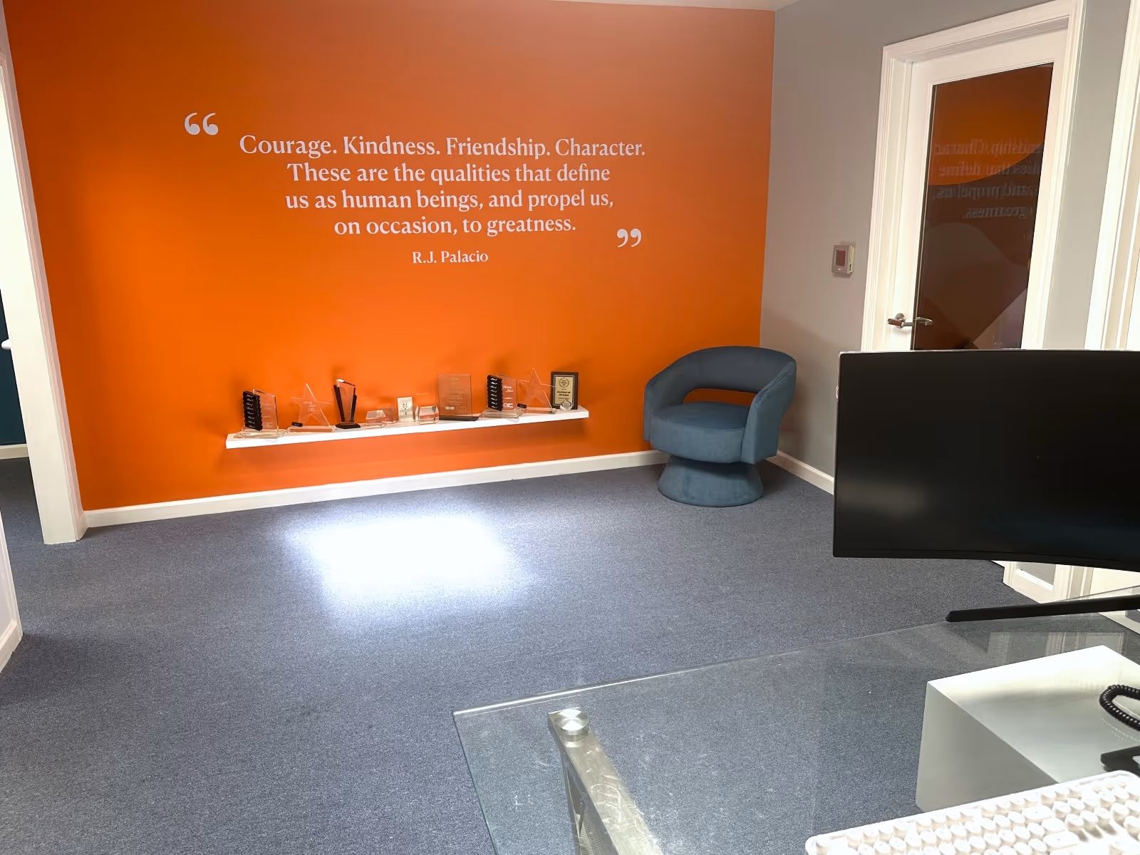 Reception area featuring an orange wall with a quote, awards shelf, and blue chair.