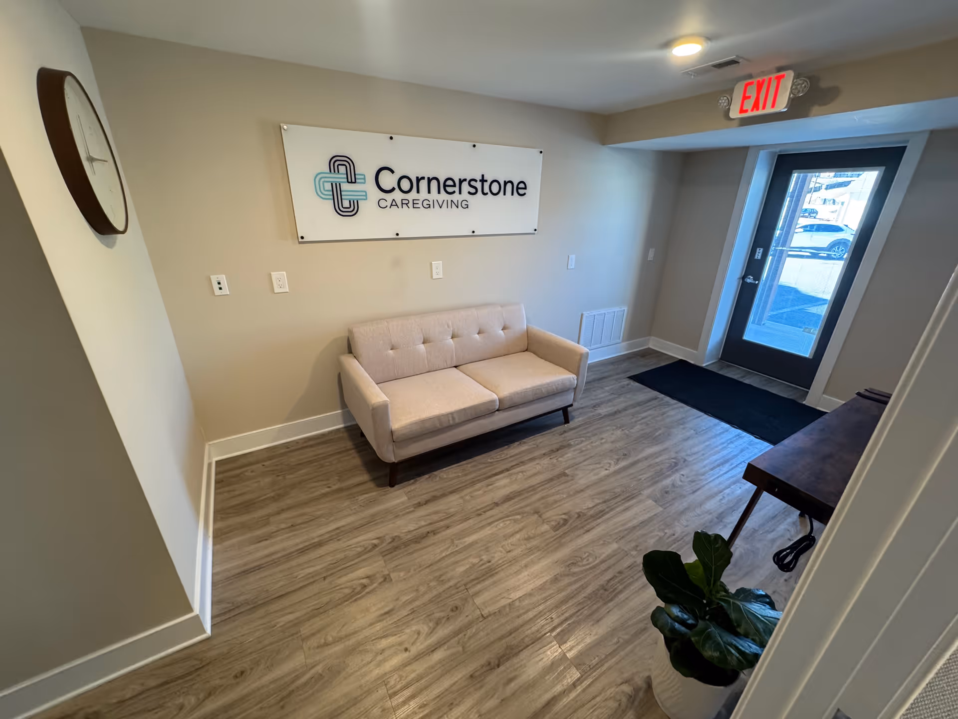 Lobby with beige sofa, Cornerstone Caregiving sign, clock, exit doorway, and potted plant.