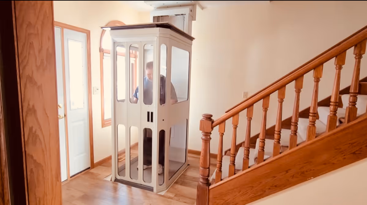 A person stands inside a small residential elevator cabin in a hallway beside a staircase.