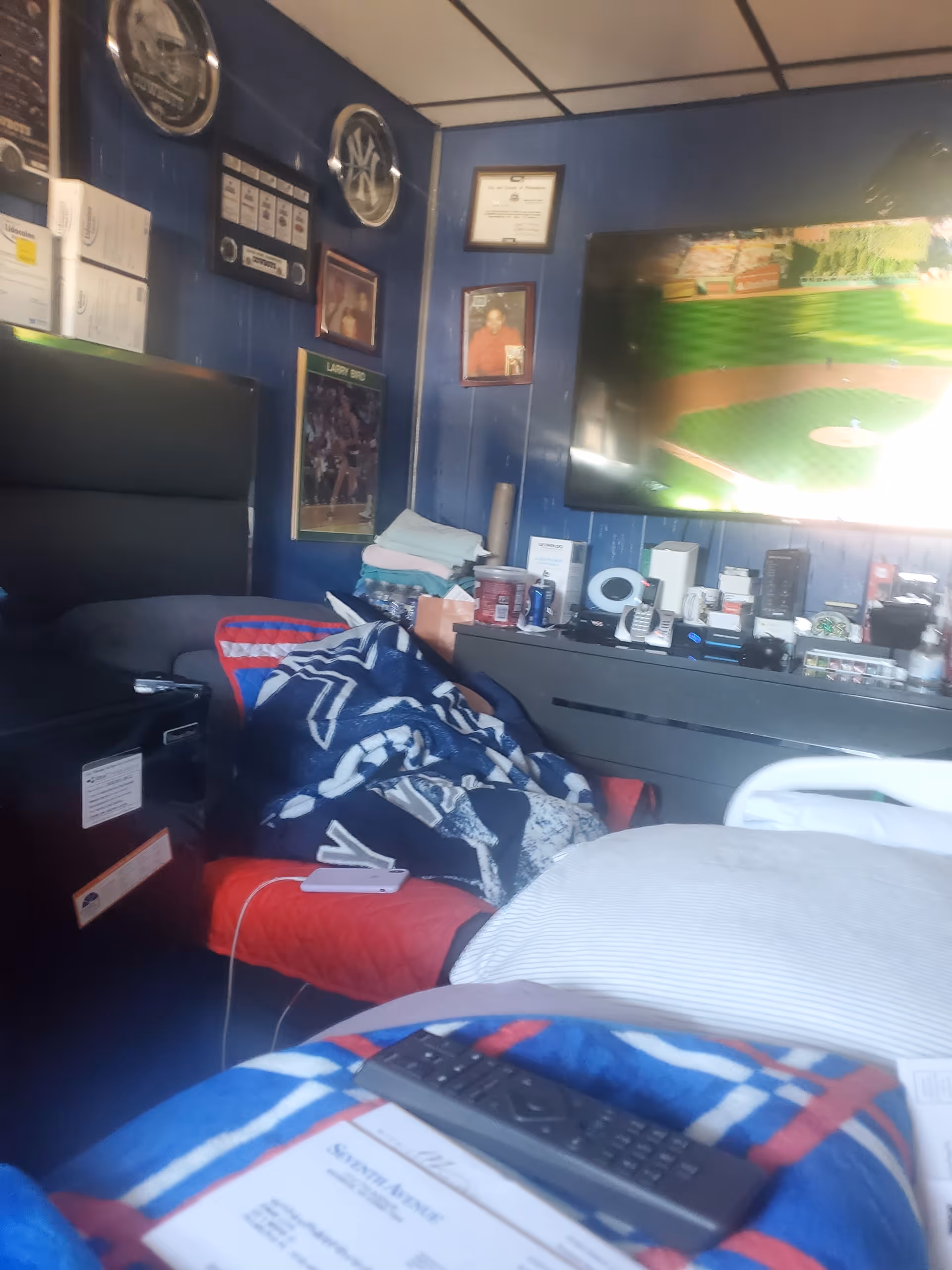 Blue-walled resident's bedroom with a bed, blankets, dresser, and mounted TV.