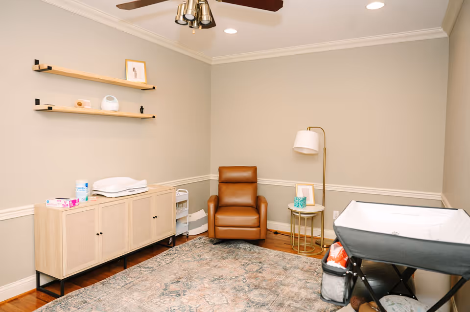 A cozy nursing room with a brown leather chair, lamp, shelves, and a bassinet.