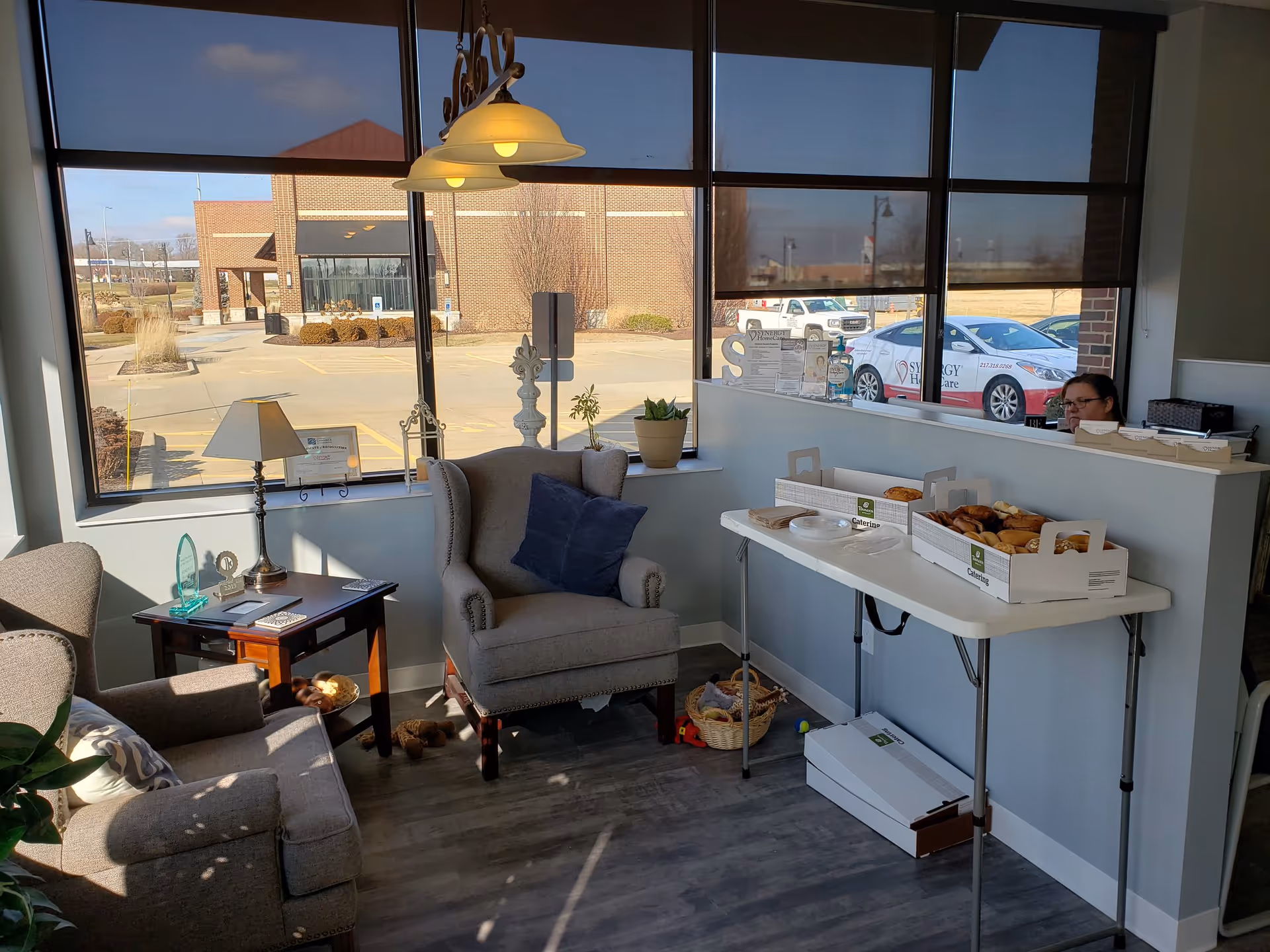 Sunlit lounge area with comfortable chairs, a pastry table, and a receptionist.