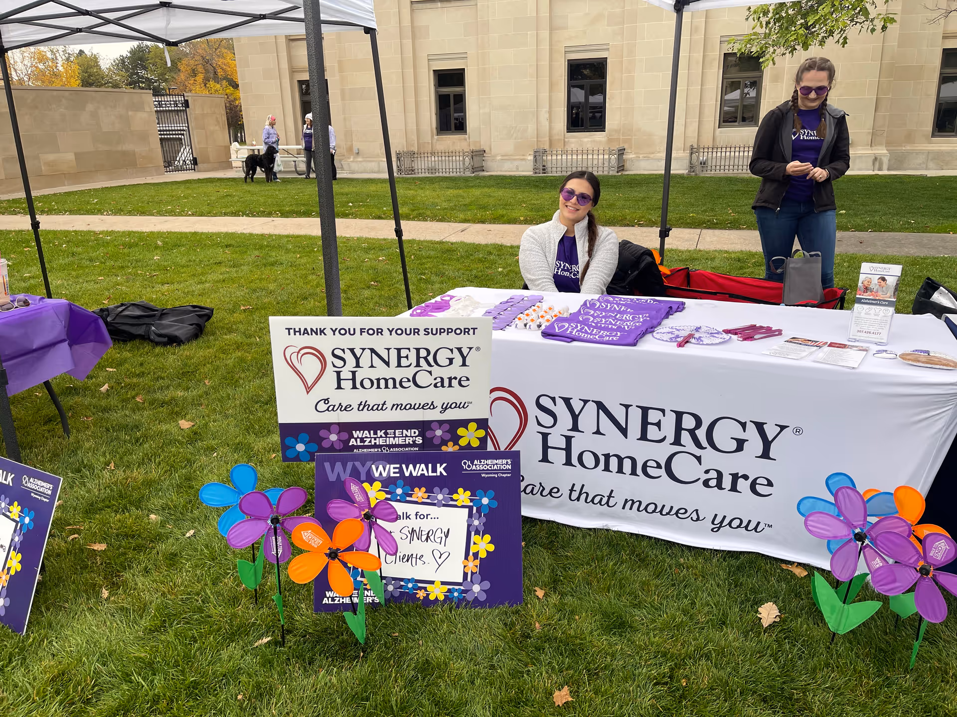 Outdoor Synergy HomeCare booth with volunteers, sign, and purple promotional materials on a grassy lawn.