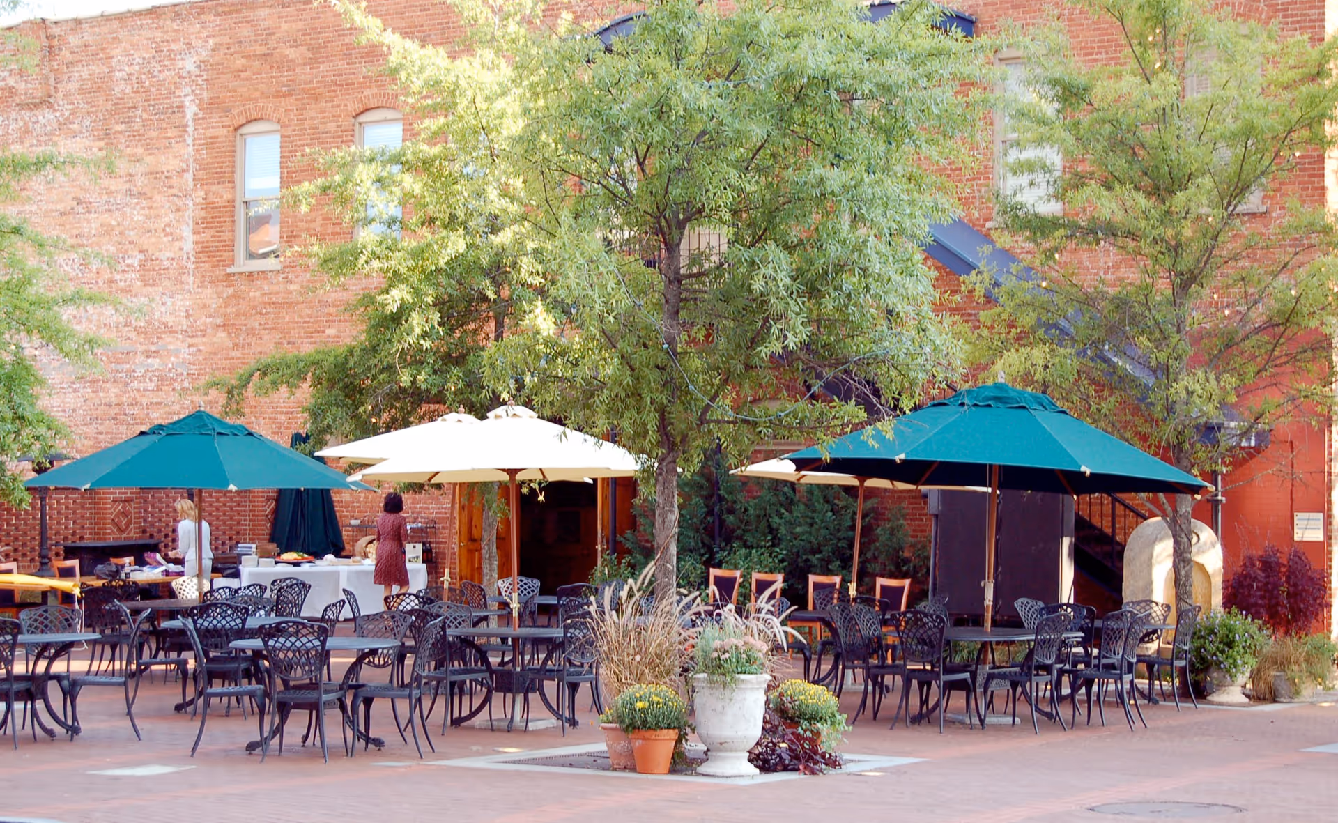 Outdoor patio with numerous black metal tables and chairs under umbrellas beside a brick building.