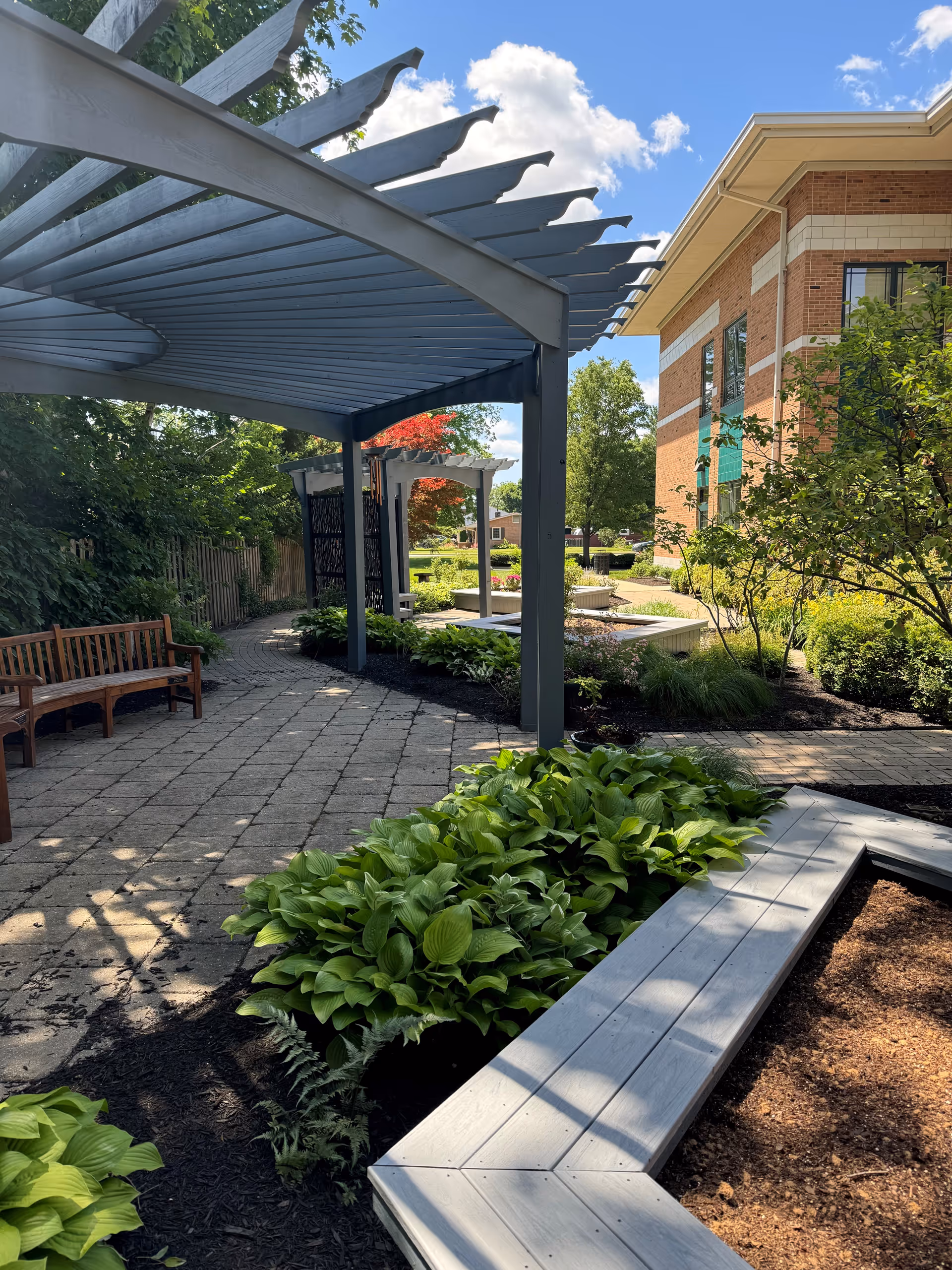 Outdoor courtyard with benches, a metal pergola, and landscaped plants beside a brick building.