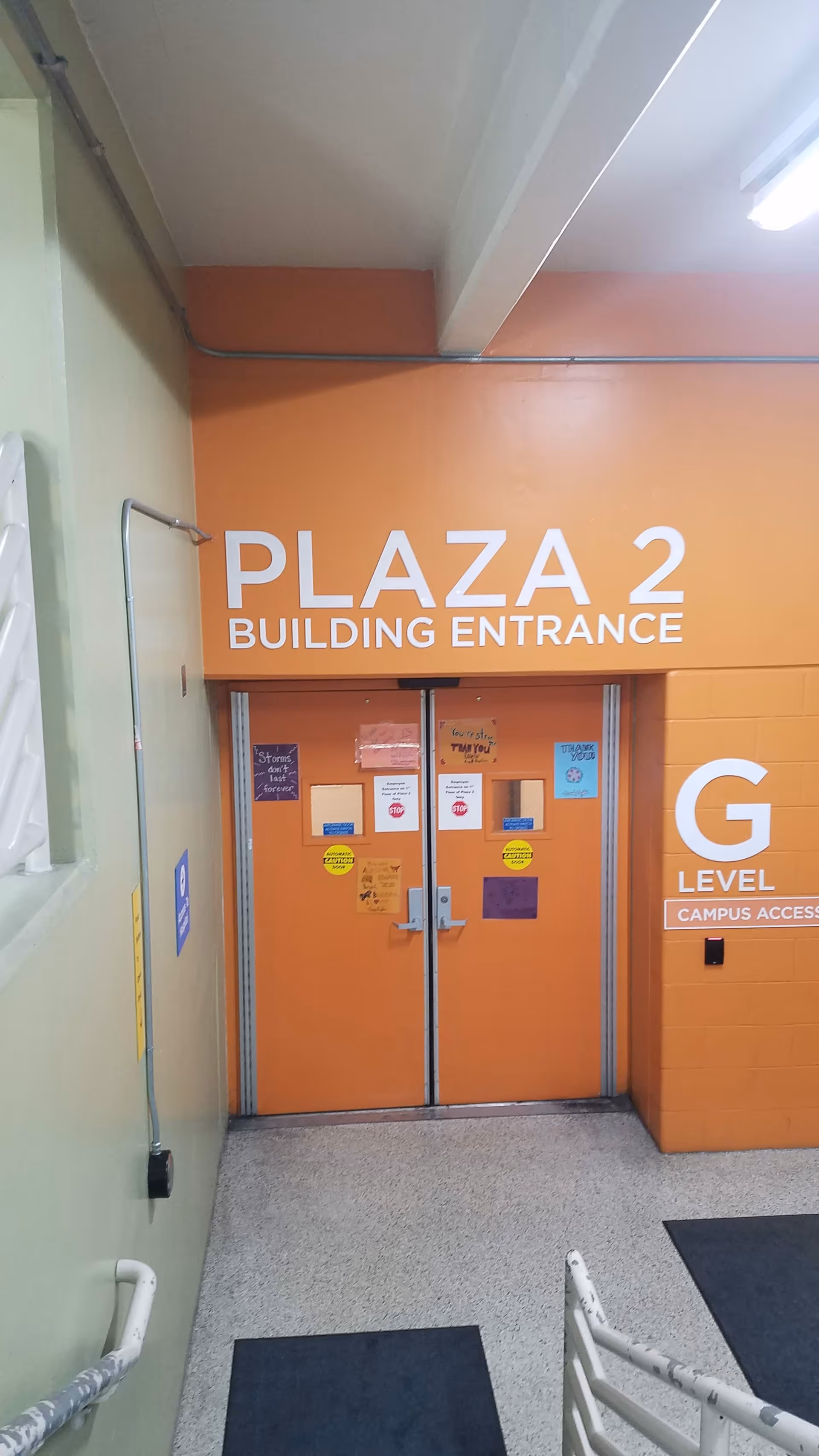 Orange double doors labeled Plaza 2 Building Entrance in a green-walled interior hallway.