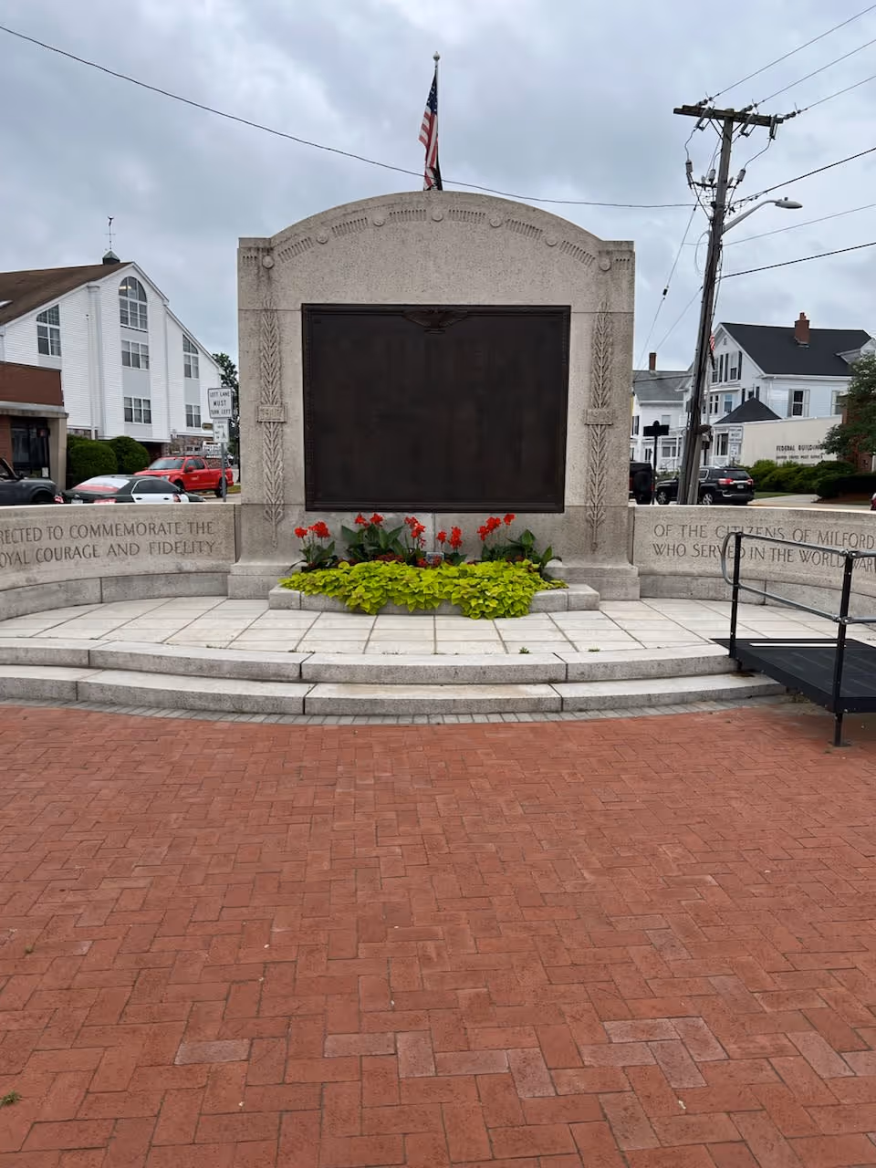 Outdoor stone monument with a dark plaque, American flag, flowers, and brick plaza.