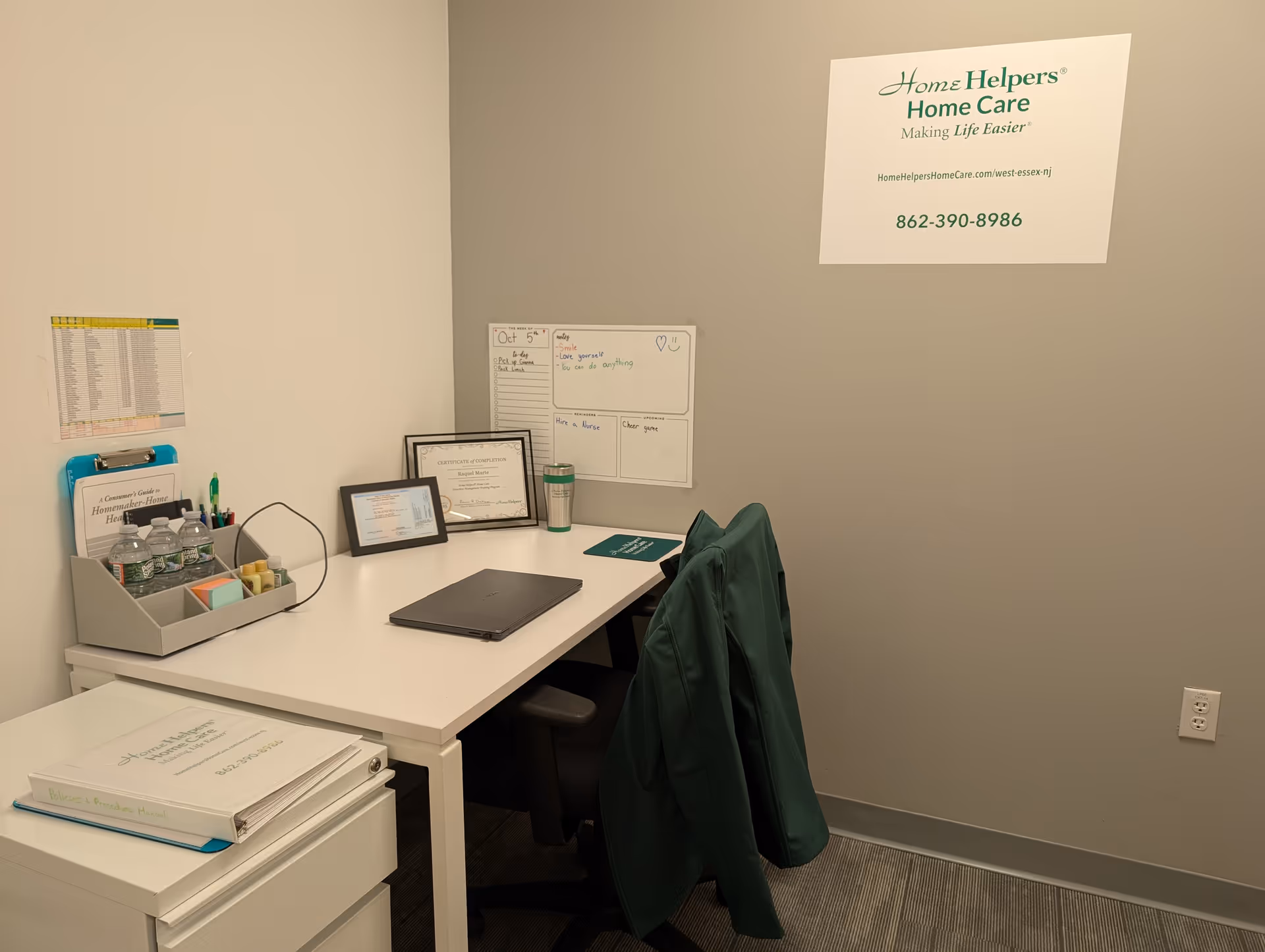 A clean office workspace with a desk, laptop, and Home Helpers sign on wall.