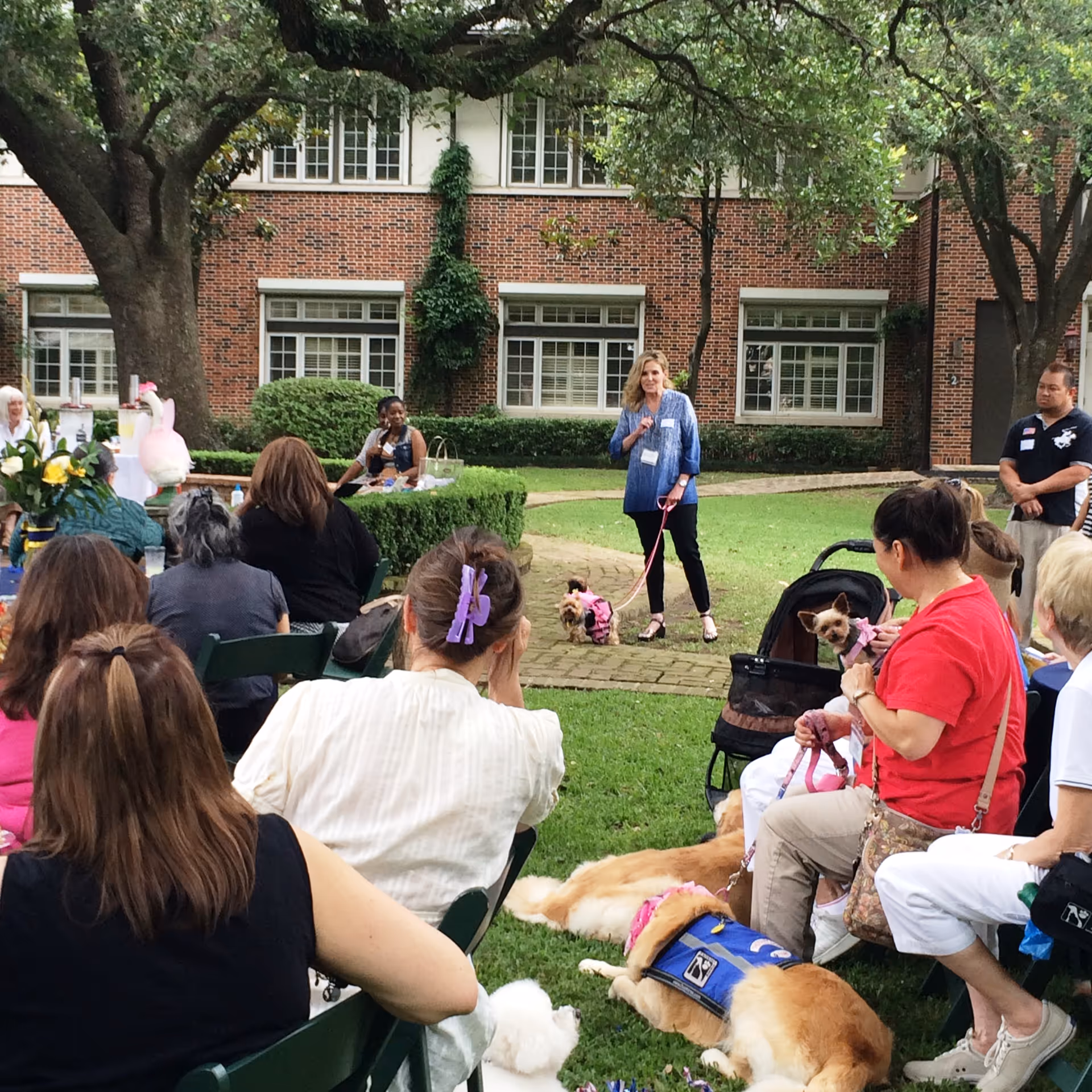 Outdoor courtyard with attendees seated, a speaker addressing the crowd, and dogs nearby.