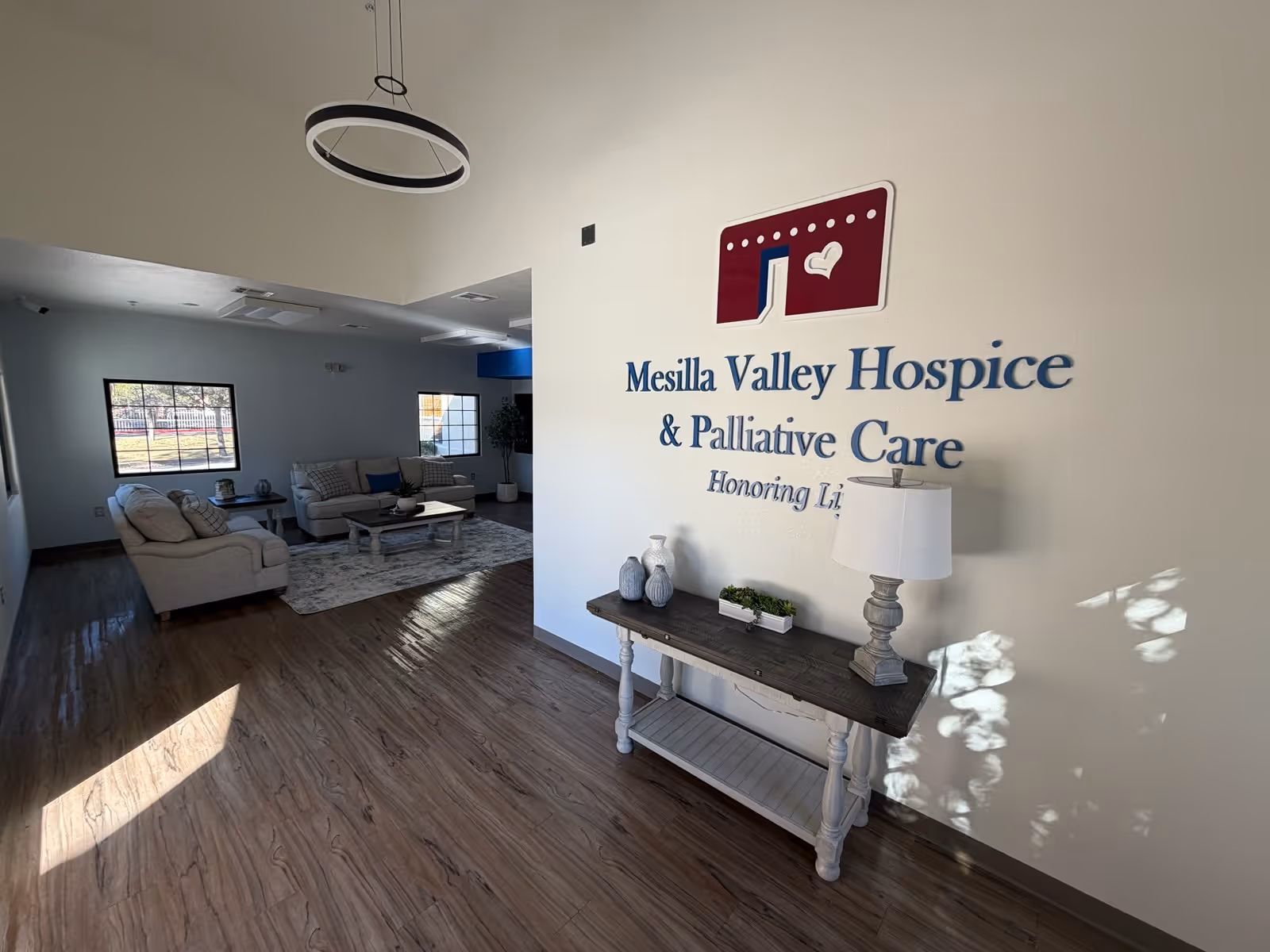 Hospice common area with sofas, coffee table, rug, and a wall sign reading Mesilla Valley Hospice.