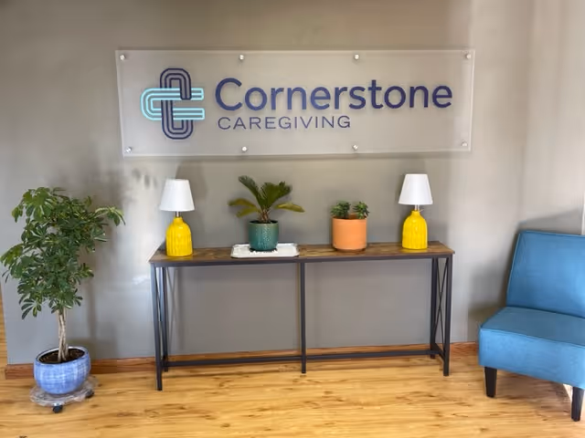 Interior lobby area with Cornerstone Caregiving sign, console table, plants, lamps, and a blue chair.