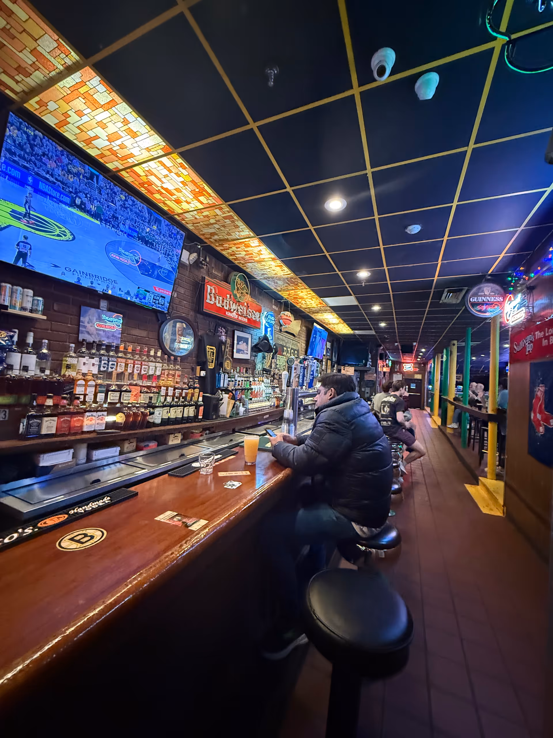 Indoor bar area with patrons at a long wooden counter and multiple TVs