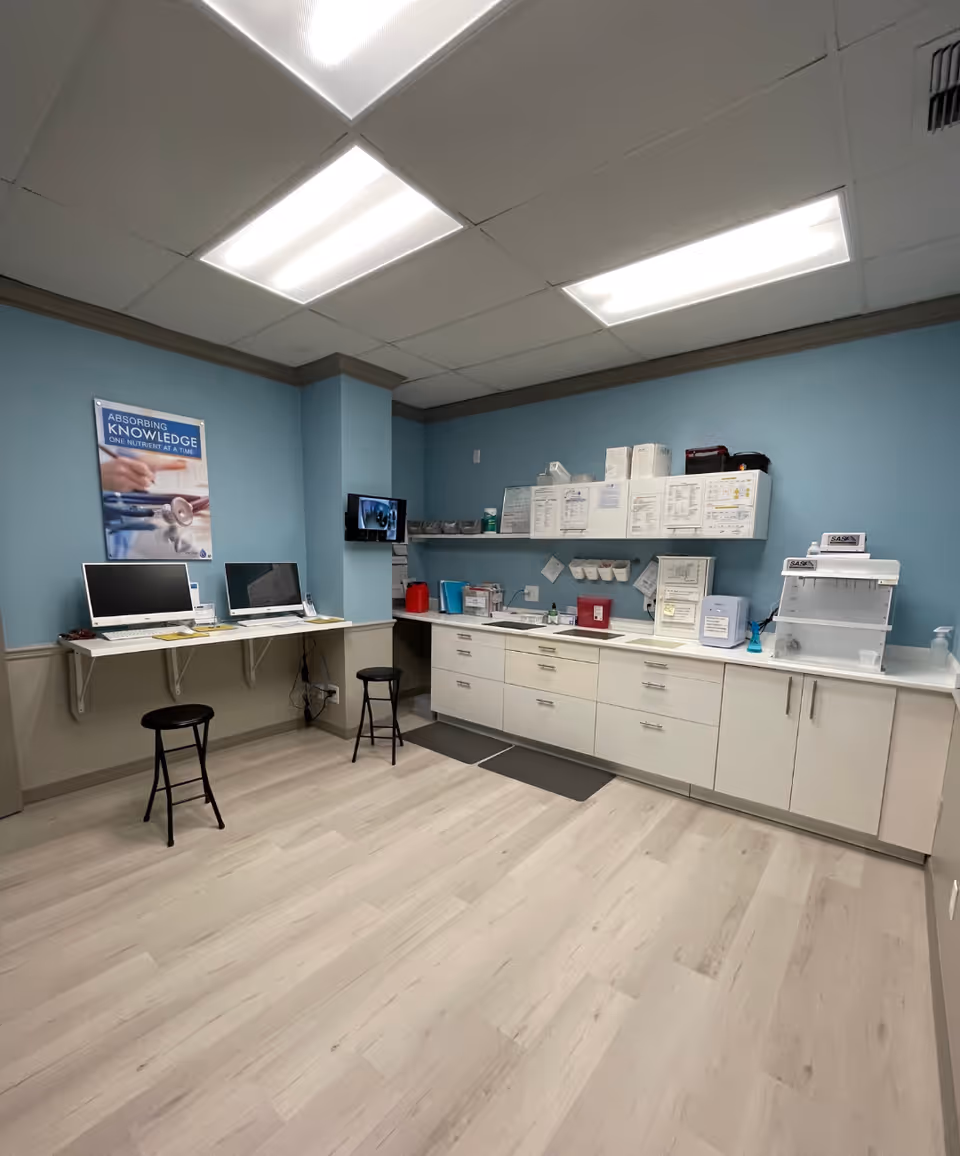 Blue-walled clinical room with a long counter, cabinets, computers, and two stools.