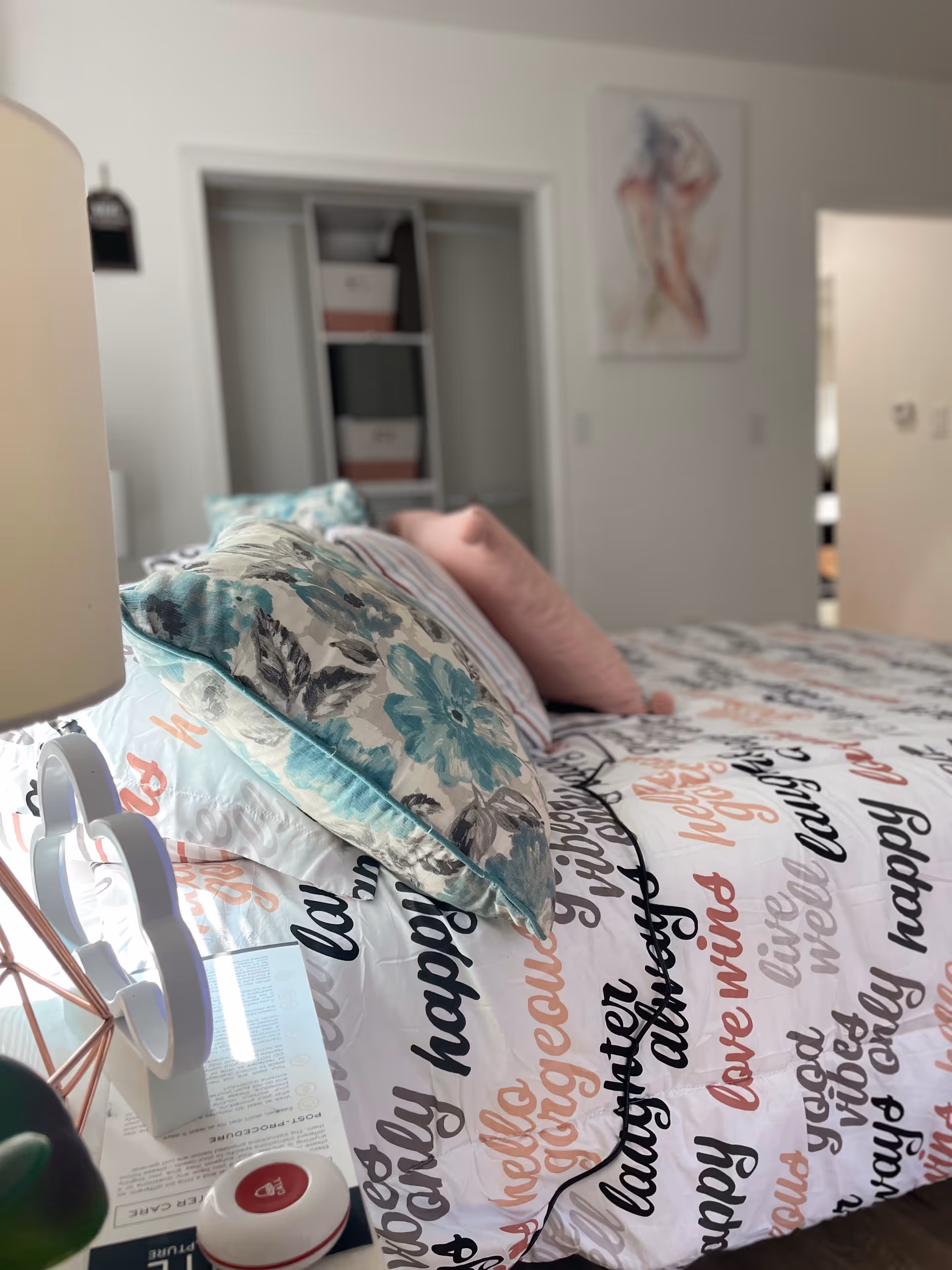 Senior living bedroom with patterned duvet, decorative pillows, lamp, nightstand, closet, and wall art.