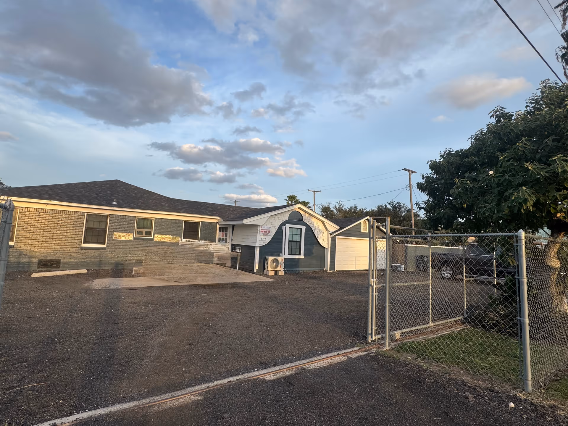 Exterior view of a single-story facility with a chain-link fence and gravel parking area.