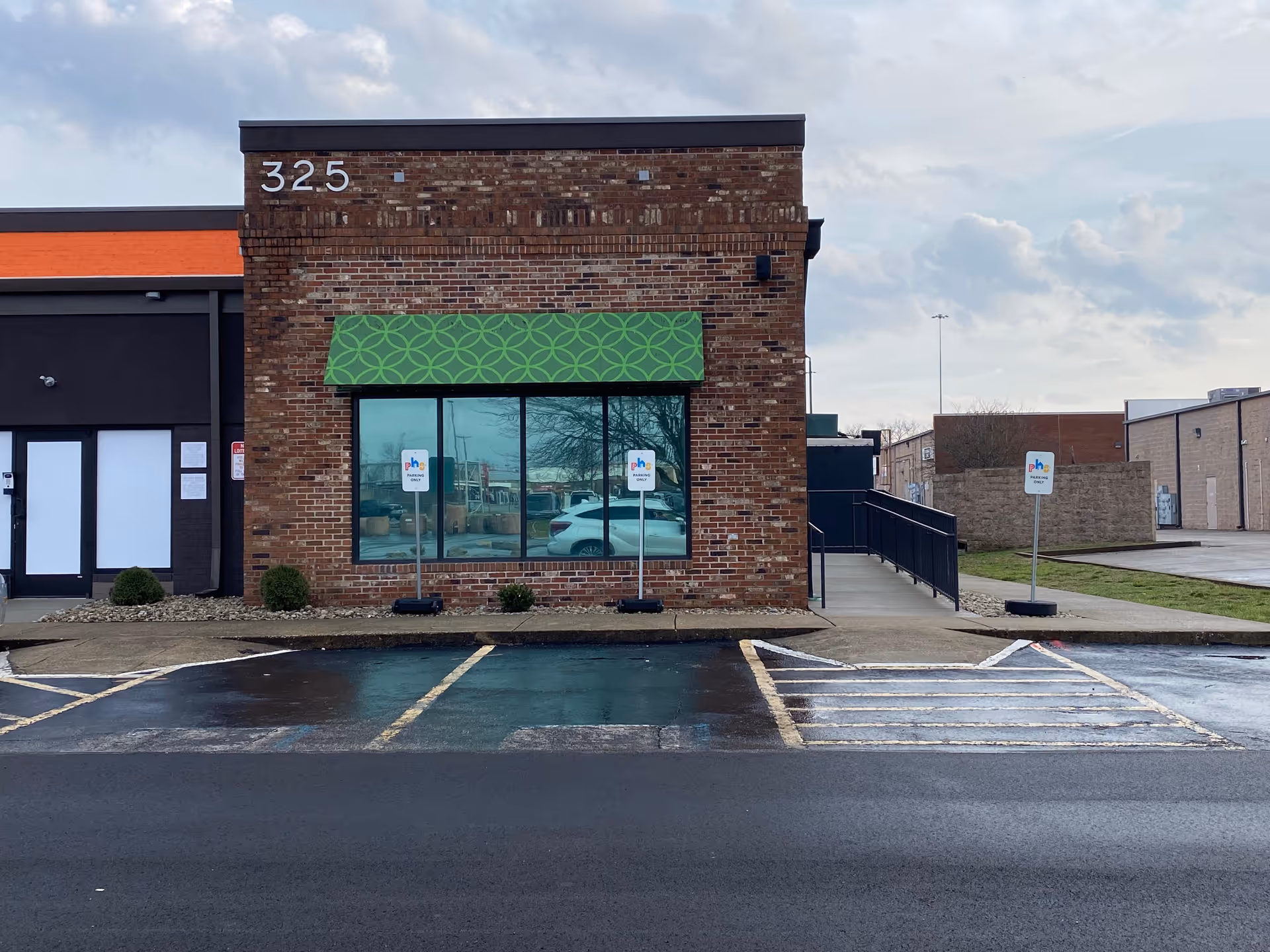 Brick medical facility with a green patterned awning, large front window, and a paved parking lot.