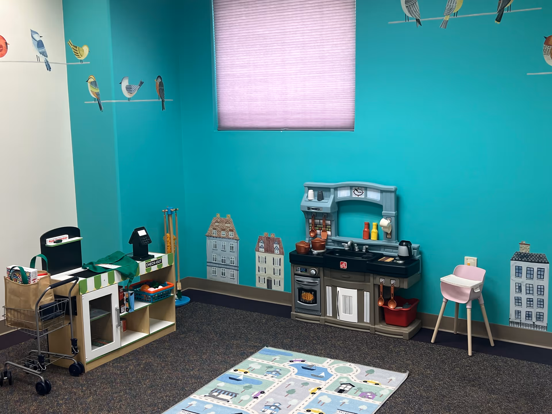 Bright teal playroom with toy kitchen, kid-sized chair, shelves, and city-themed wall murals.