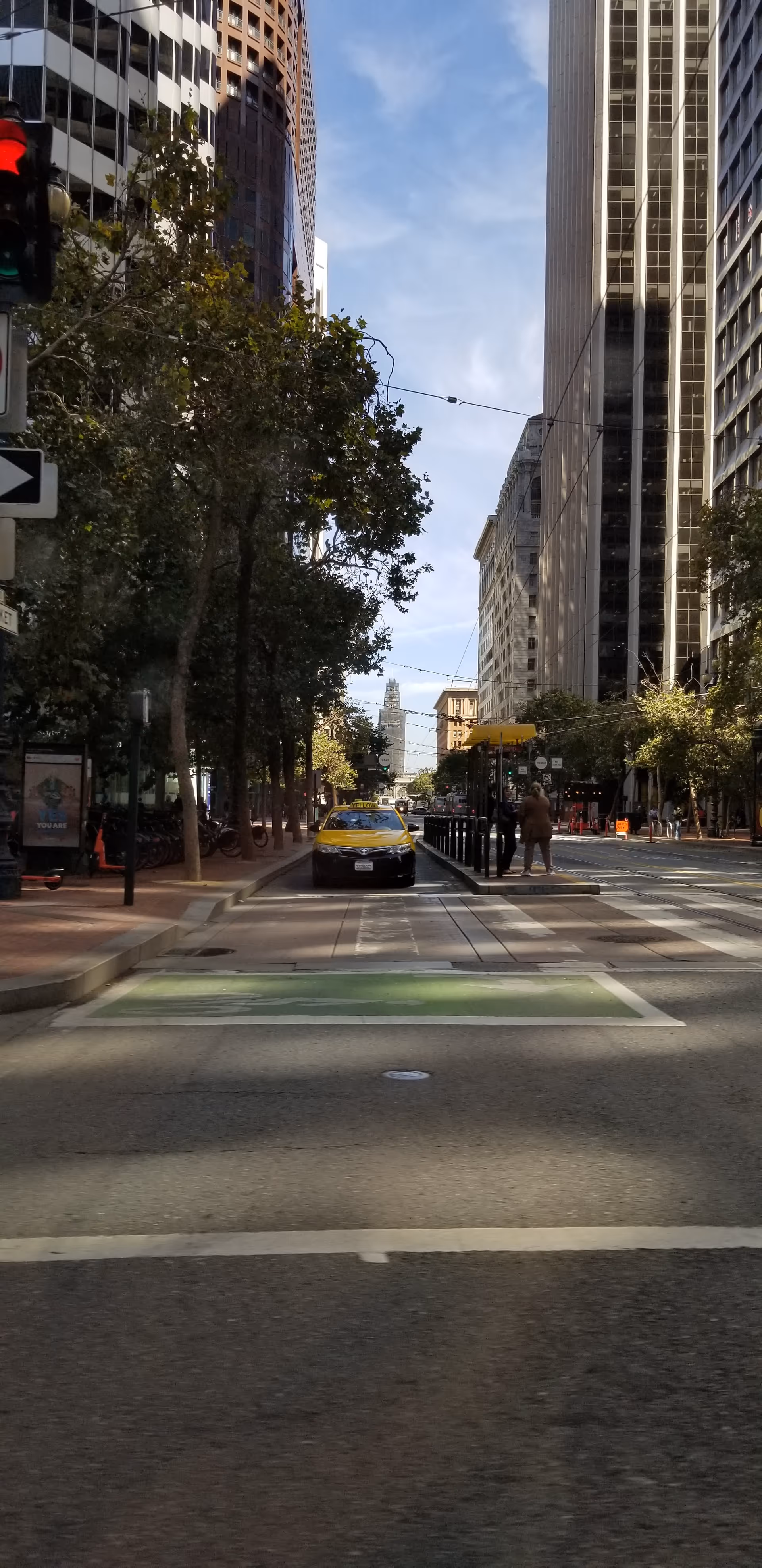 Urban street scene with tall buildings, trees, and pedestrians, including a yellow taxi in the center.