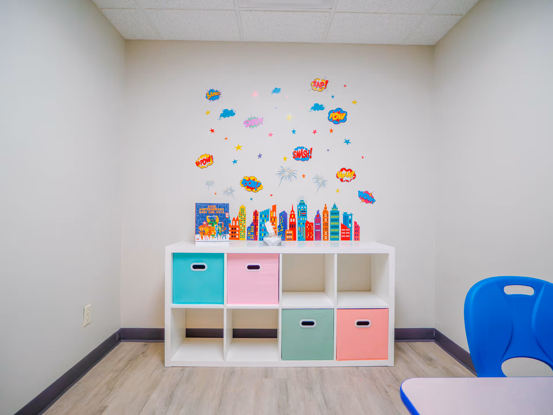 Small colorful play area with cubby storage, wall decals, and blue chair in a neutral room.