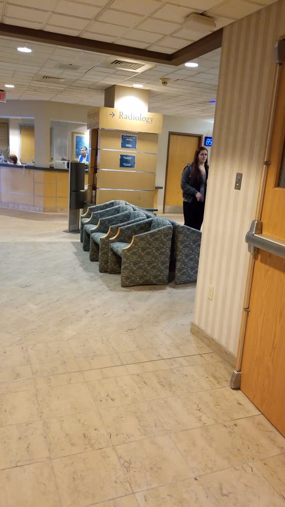 Hospital lobby with teal patterned chairs, a reception desk, and a radiology sign by a hallway.