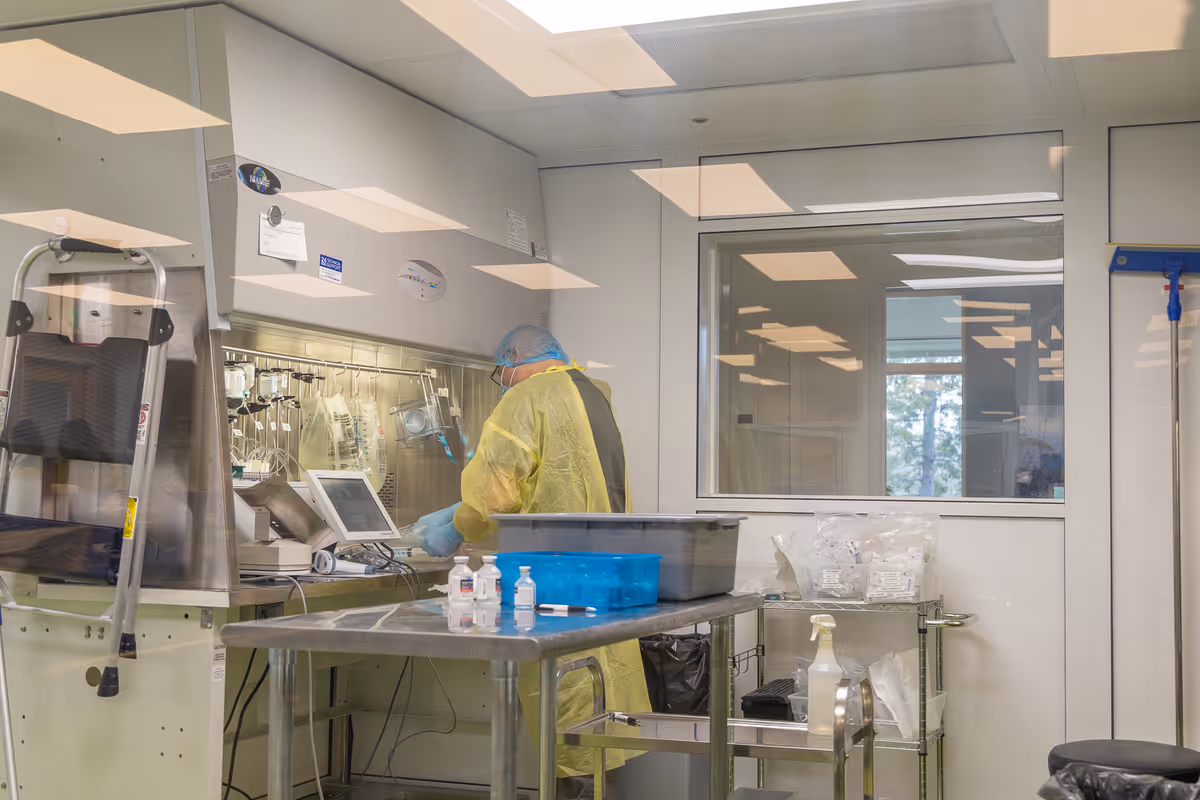 A healthcare worker in PPE works at a sterile hood in a clinical infusion prep area.