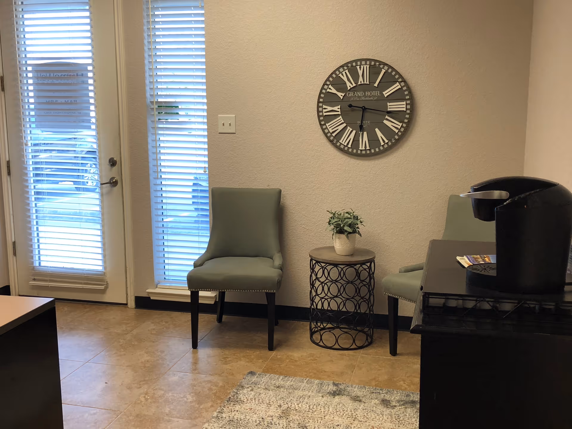 A lobby seating area with two mint-green chairs, a round metal table, clock, and coffee station.