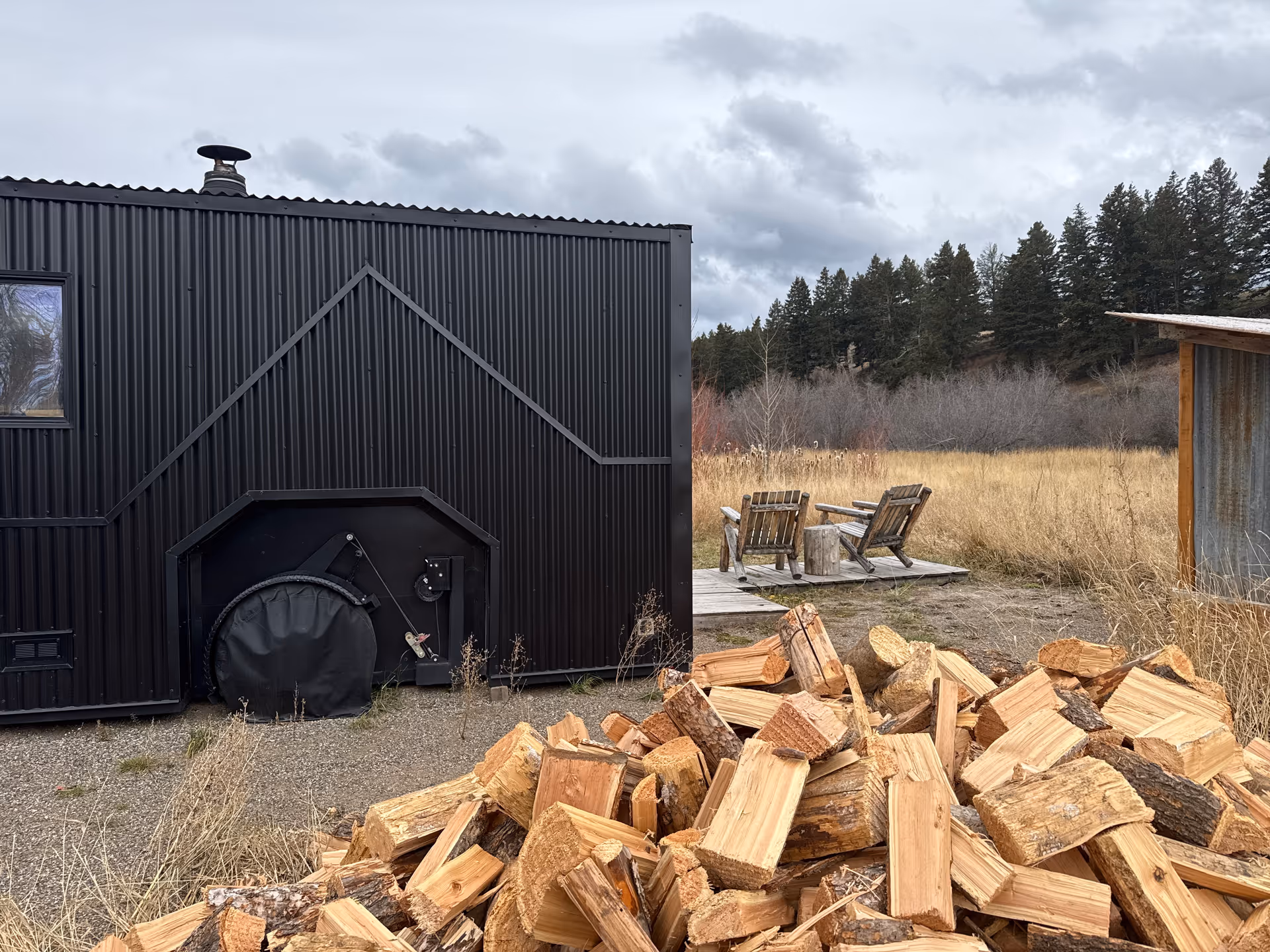 A dark, modern exterior building with a wooden deck, chairs, and a pile of firewood.