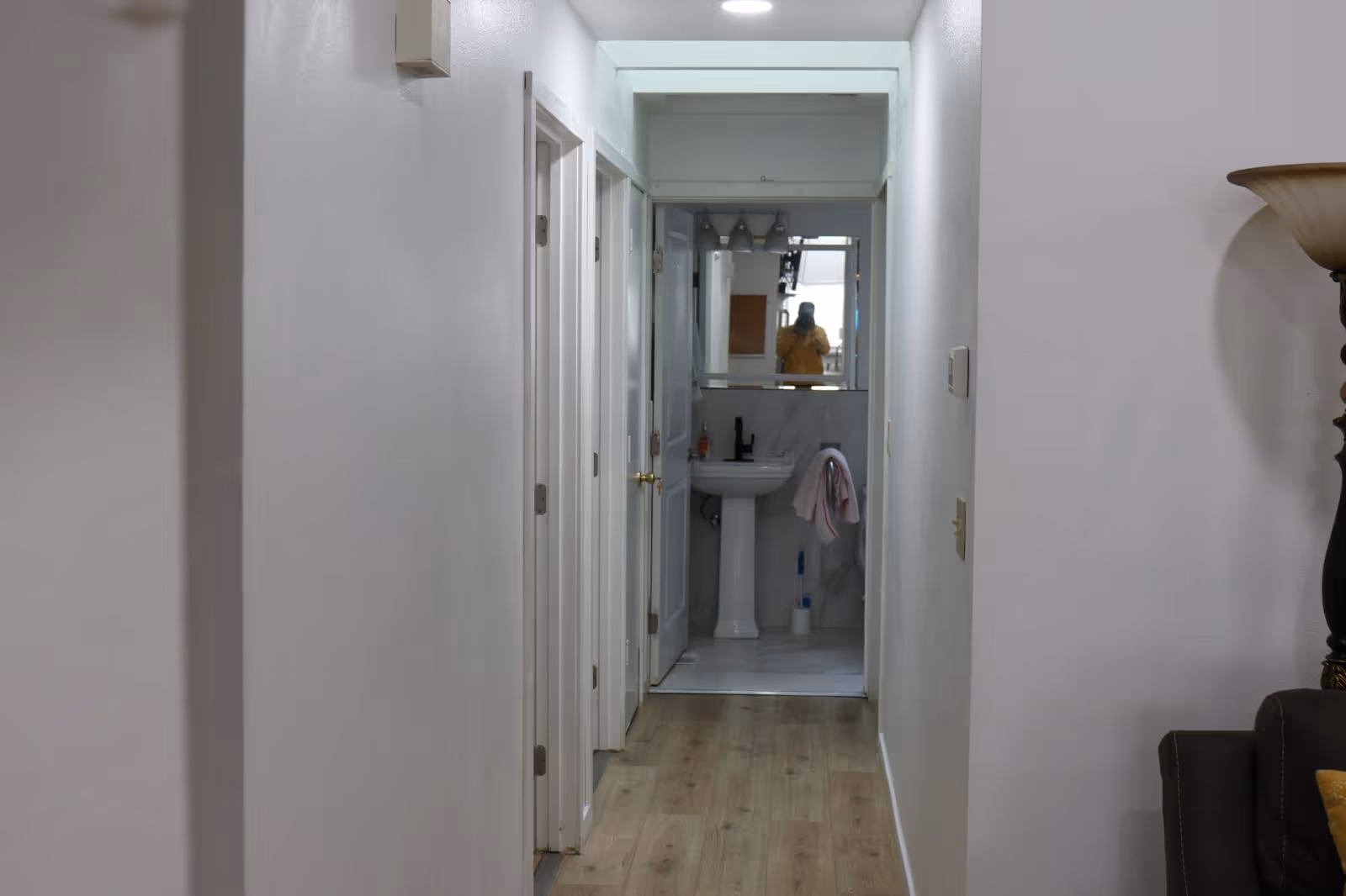 White hallway leads to a bathroom with pedestal sink and mirror.