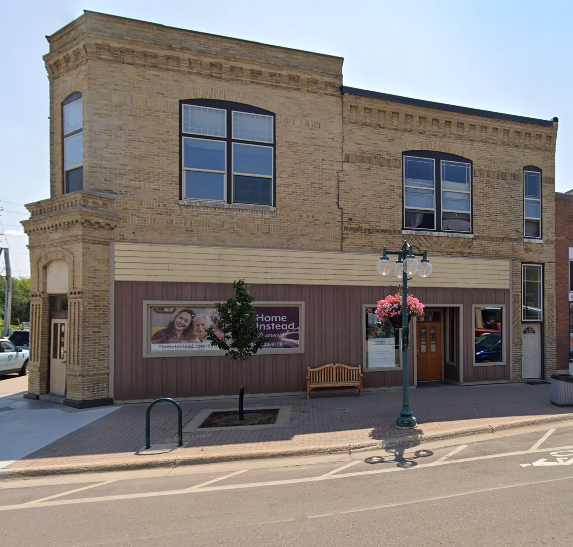 Exterior view of a two-story brick building with a Home Instead storefront, bench, tree, and streetlamp.