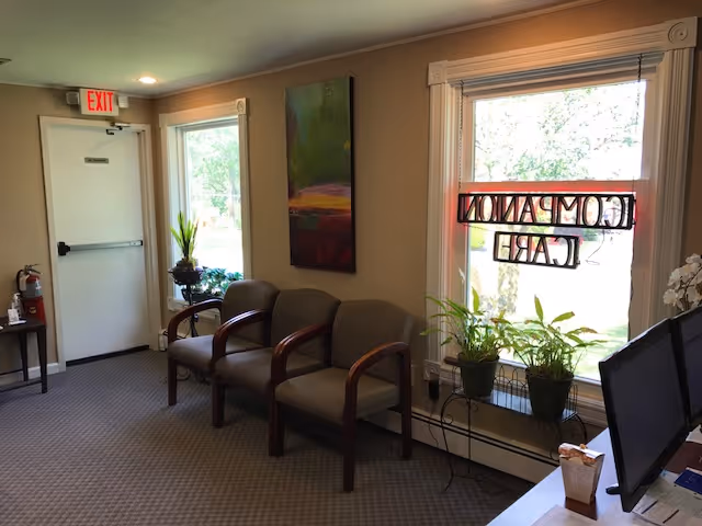 A small waiting area with brown upholstered chairs, plants, and a computer near the window.